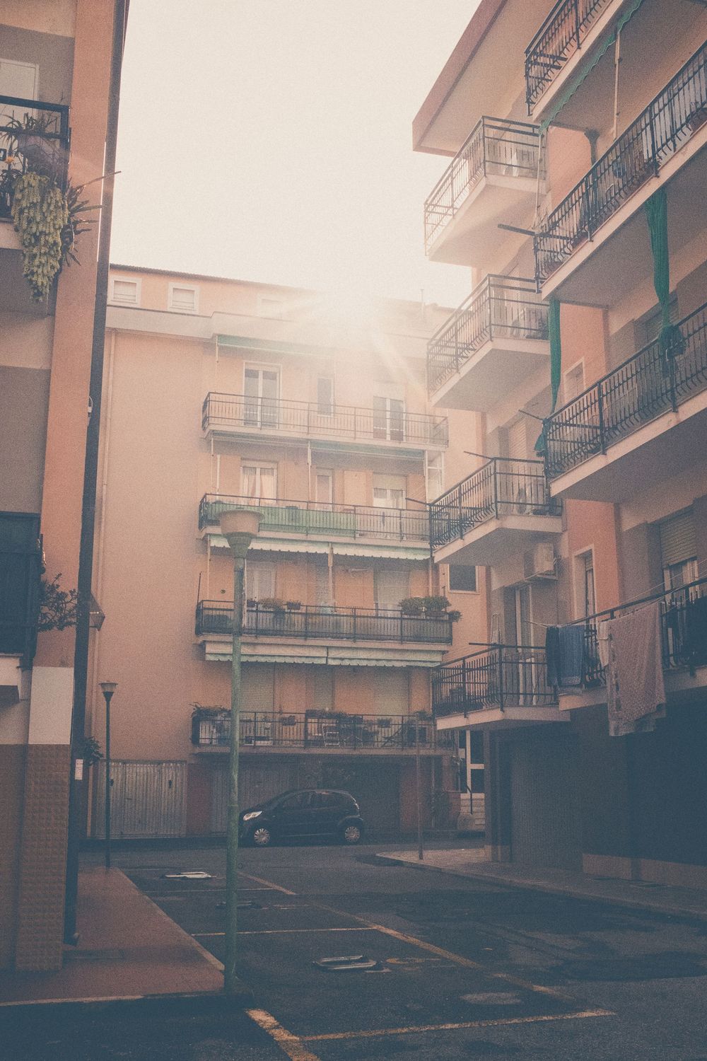 A moody urban apartment building with balconies and parking lot captured during golden hour with sun flare effect.