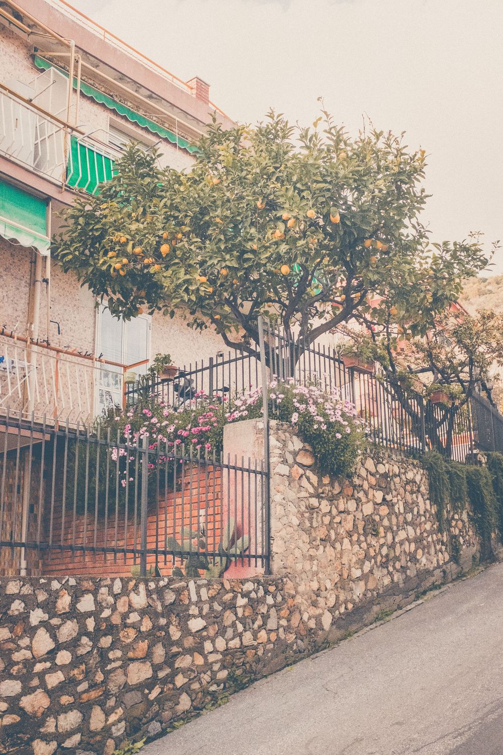 A Mediterranean stone wall with a citrus tree and green awnings on a residential building along a sloping street.