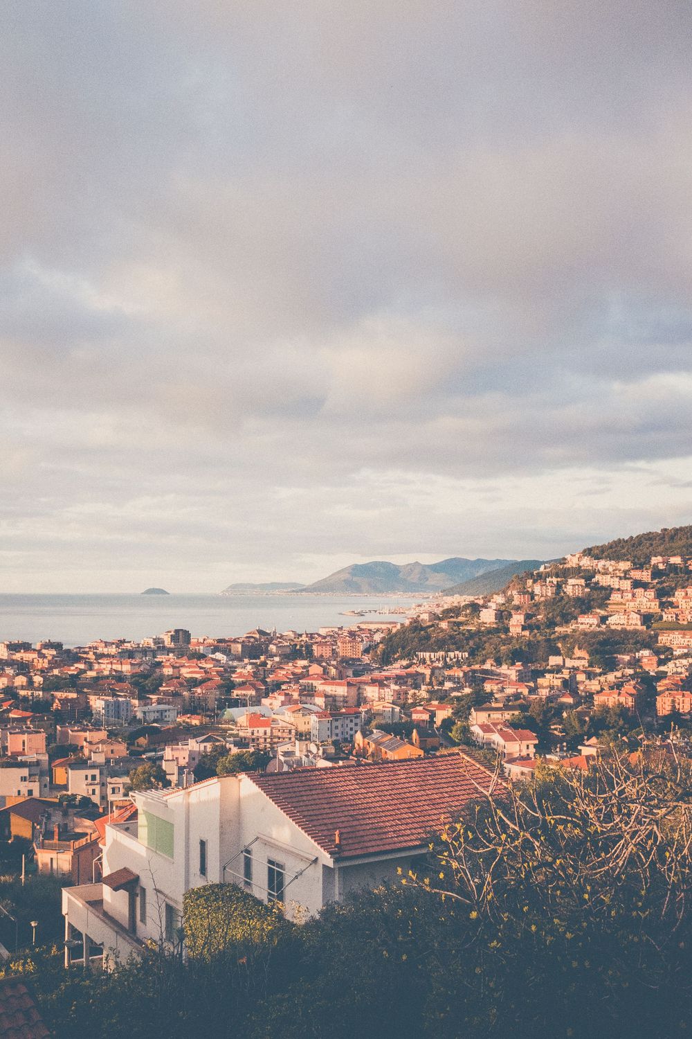 Coastal Mediterranean town with terracotta roofs cascading down hillside at sunset, overlooking misty ocean and mountains.