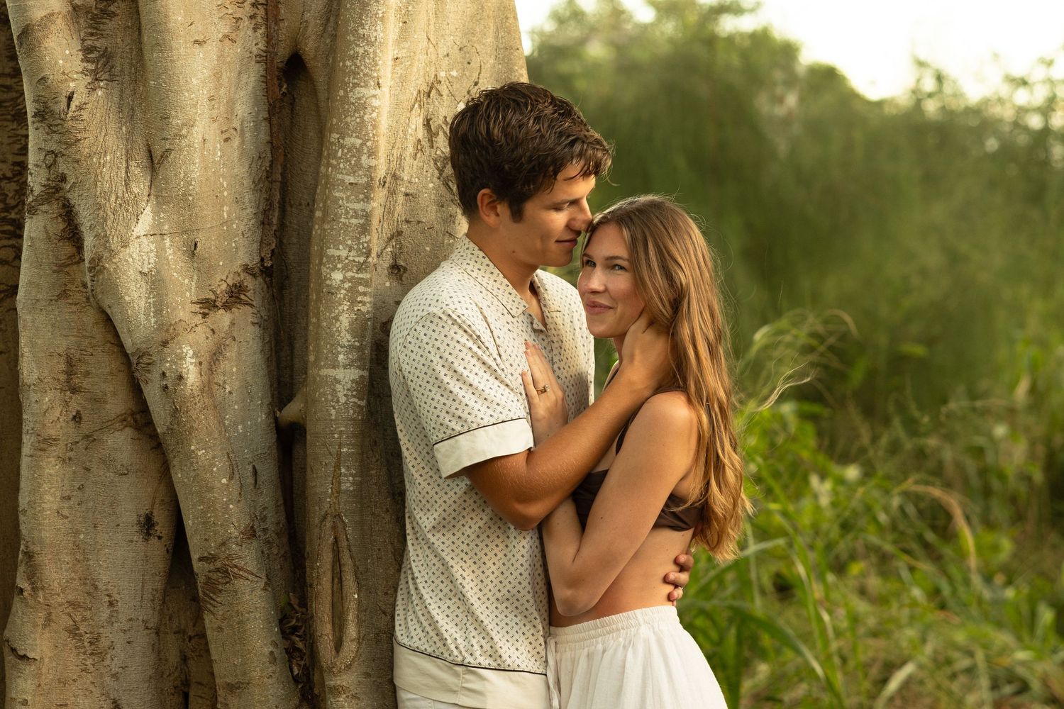 A romantic couple shares an intimate embrace against a large tree trunk in warm golden sunlight.