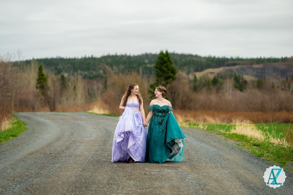 Two people in formal dresses walking together on a rural road surrounded by green fields and forests.