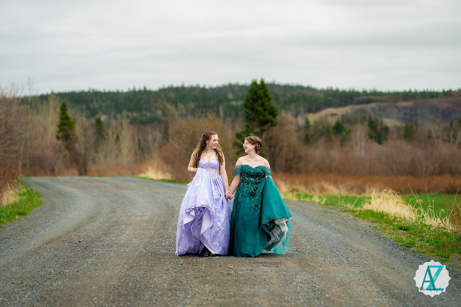 Two people in formal dresses walking together on a rural road surrounded by green fields and forests.
