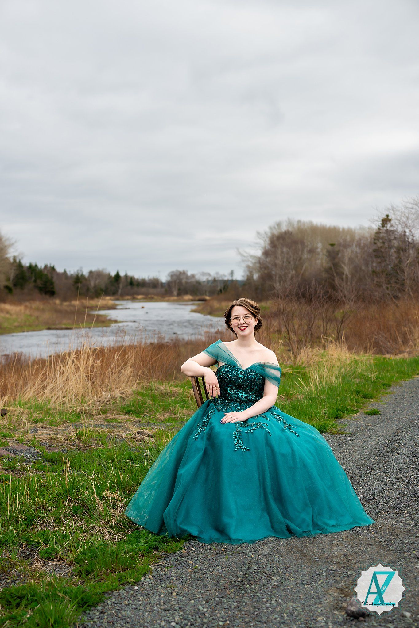Elegant teal ball gown with off-shoulder design photographed on gravel path near river and autumn landscape.