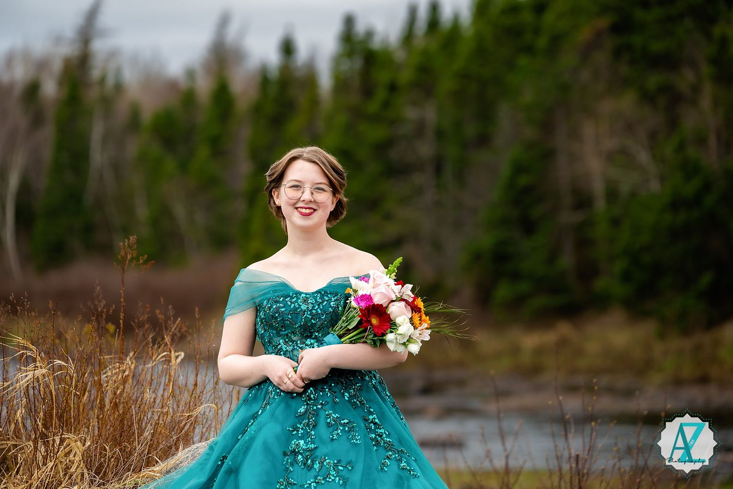 Someone wearing a teal formal gown holds a colorful bouquet while standing near a lake with evergreen trees in background.