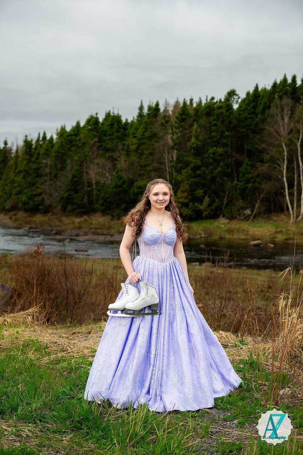 A person in a flowing lavender gown holds ice skates while posing near a lake surrounded by evergreen trees.