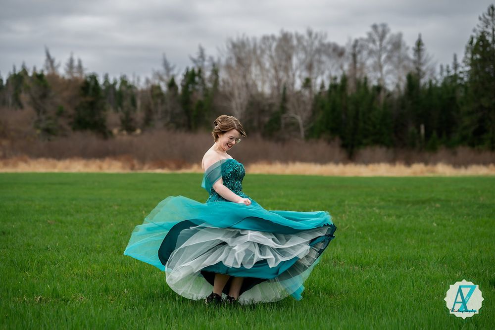 A person in a flowing turquoise dress twirls gracefully in a green field with trees in the background.