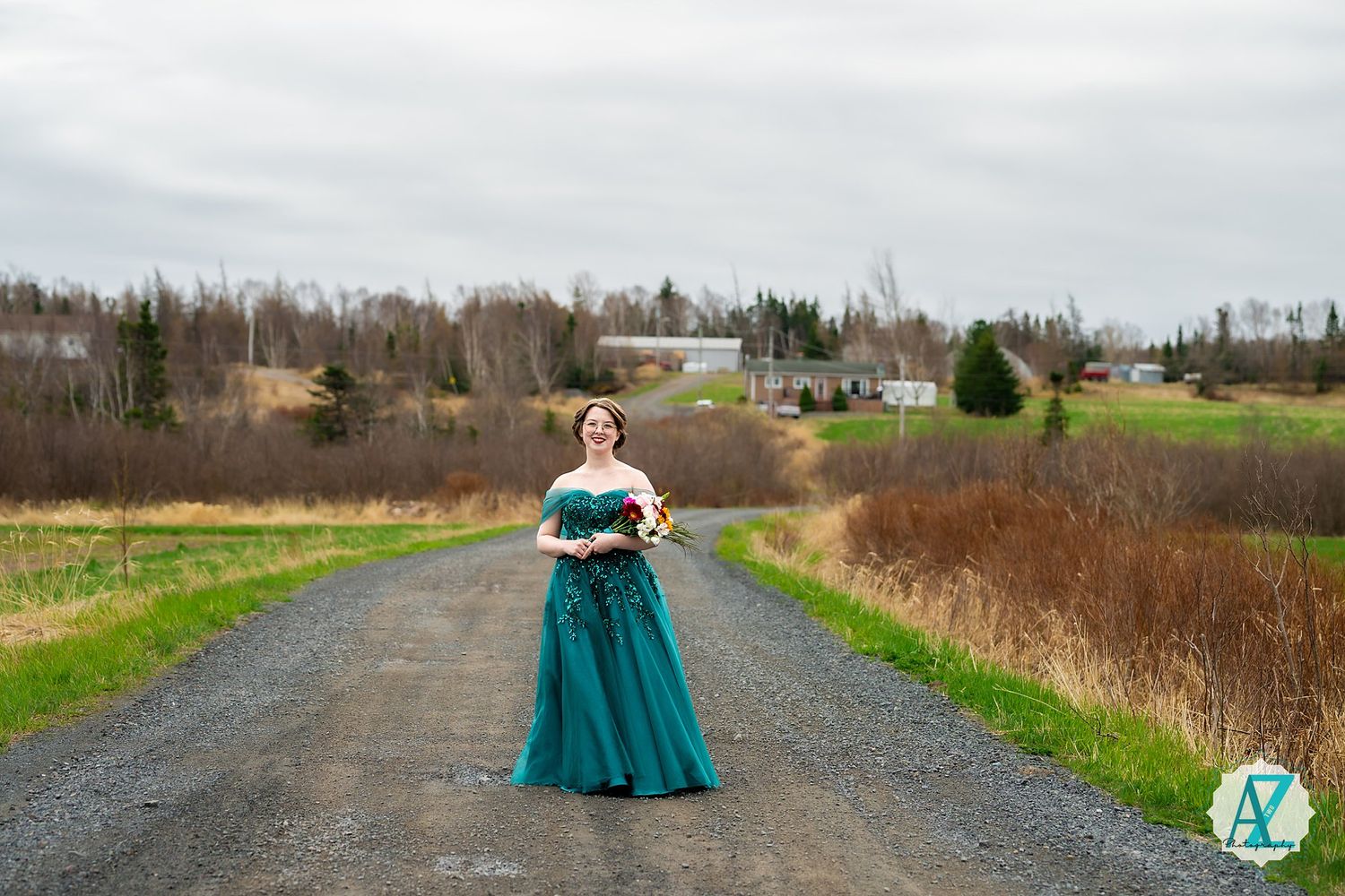 Someone in an elegant teal ballgown poses on a rural gravel road surrounded by countryside scenery.