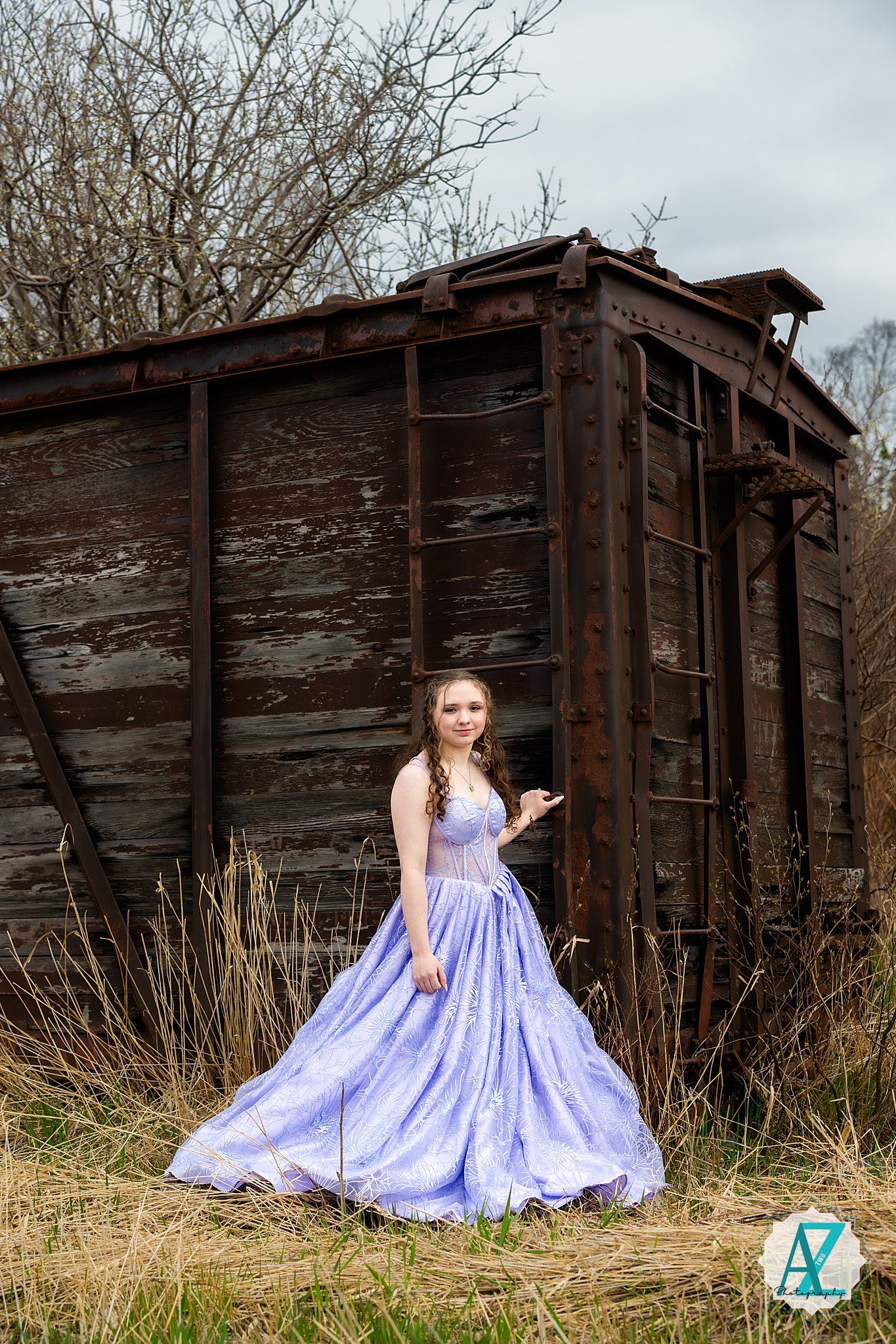 Figure in a flowing lavender ballgown poses against rustic wooden storage car backdrop in outdoor setting.