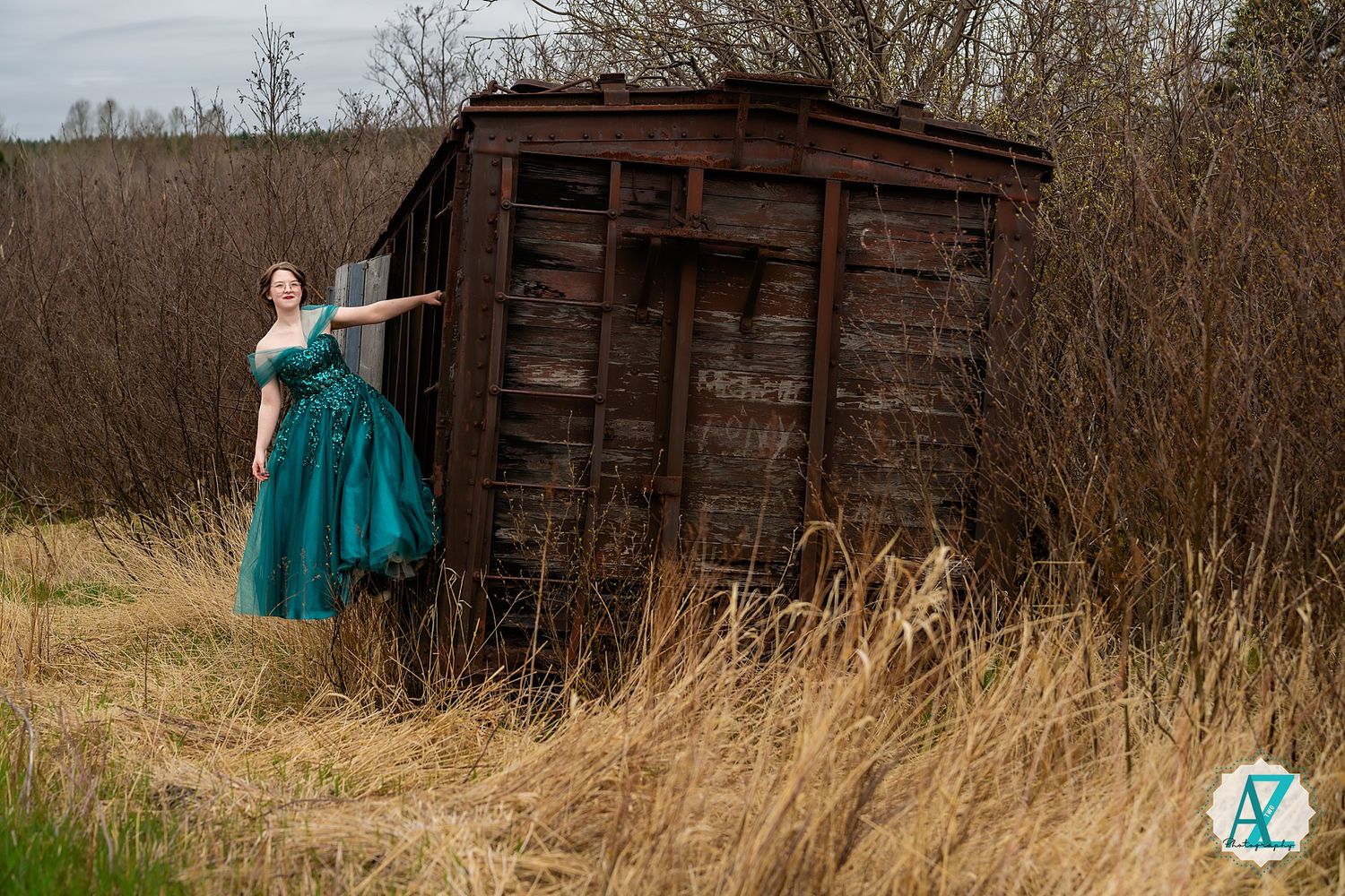 A figure in a teal vintage dress poses dramatically next to an old wooden shed in a field of tall brown grass.