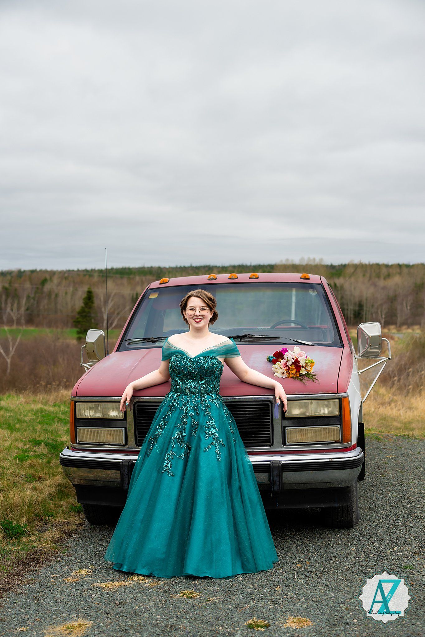 Someone in a teal ballgown poses in front of a red pickup truck on a gravel path with autumn fields in background.