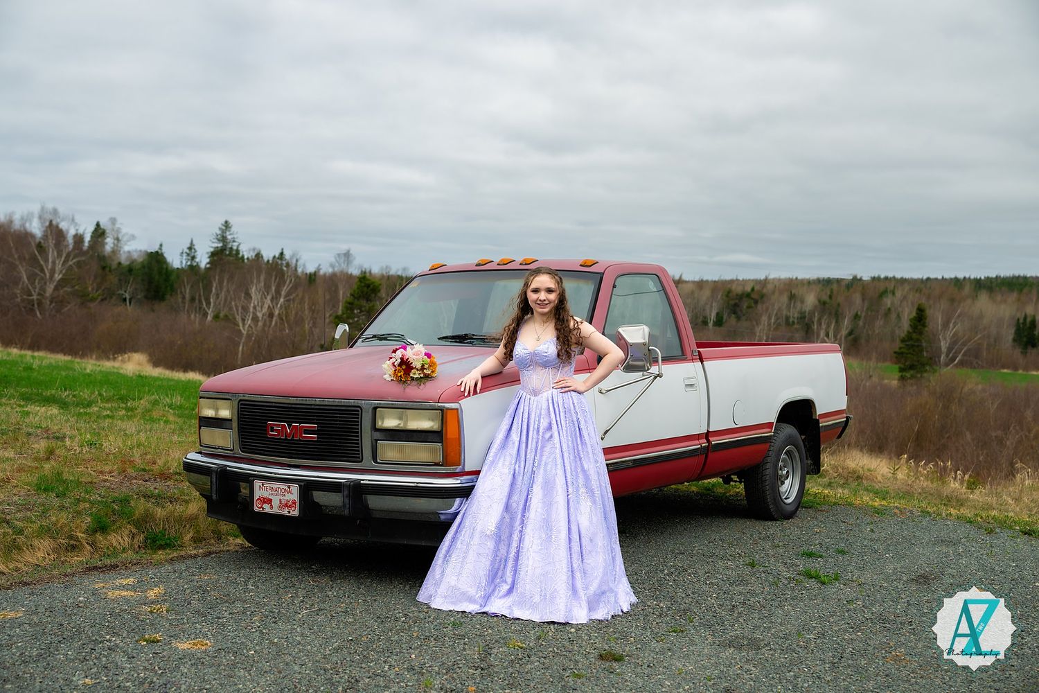 A person in a lavender prom dress poses next to a vintage red GMC Sierra pickup truck in a rural field.