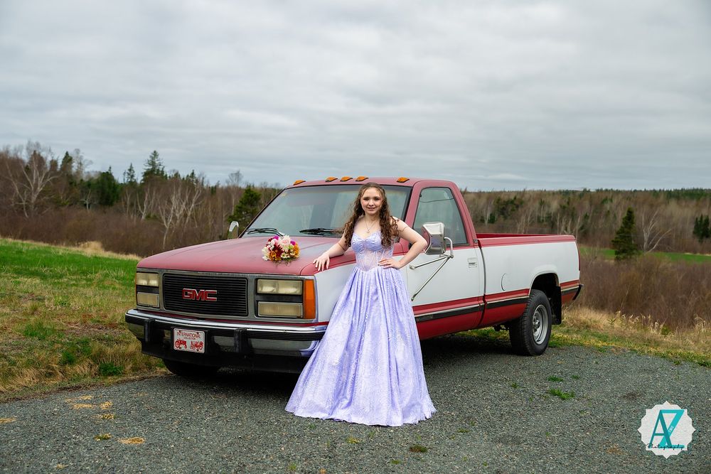 A person in a lavender prom dress poses next to a vintage red GMC Sierra pickup truck in a rural field.