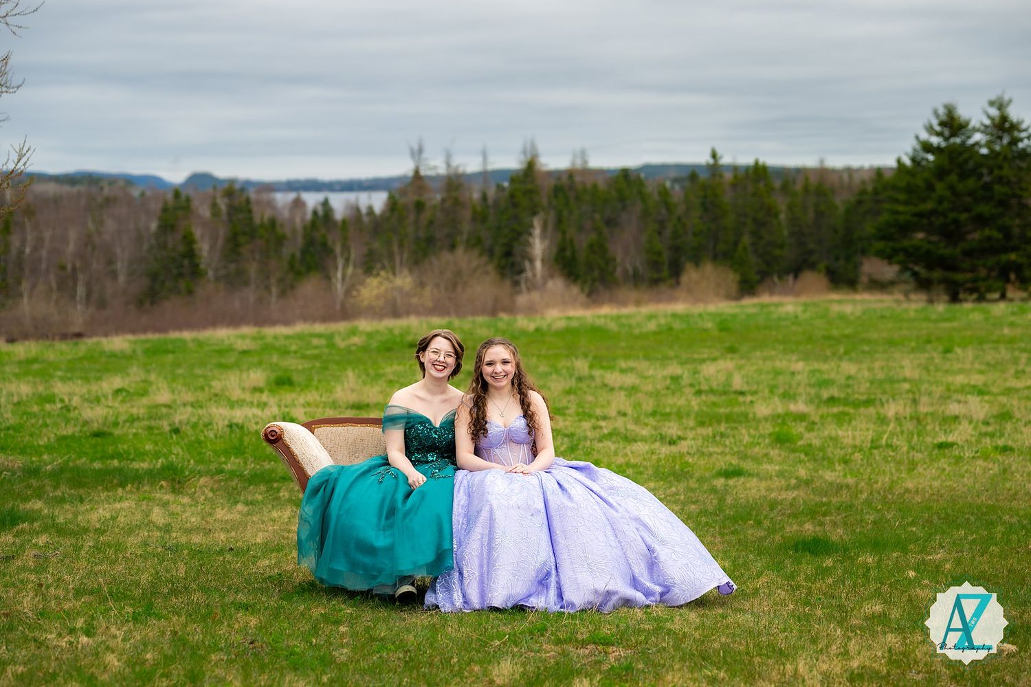 Two formal gowns in green and lavender grace a hillside meadow with evergreen forest backdrop.