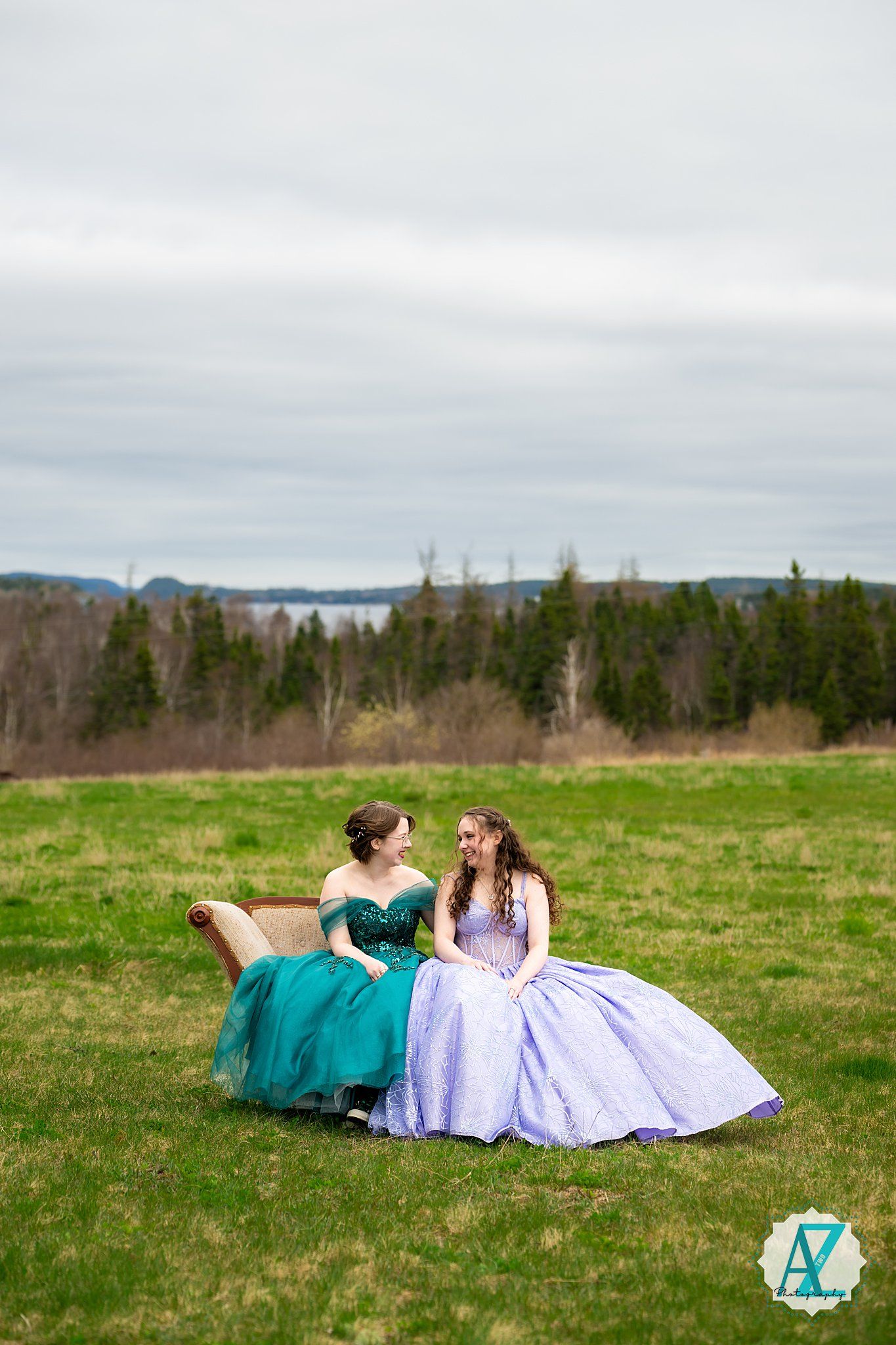 Two elegant ball gowns in teal and lavender grace a rustic outdoor bench against a scenic rural landscape.