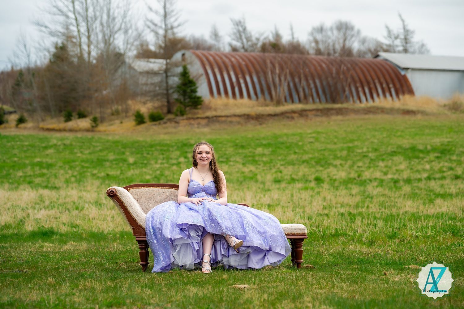 A lavender ballgown contrasts with rustic bench and quonset hut in rural landscape setting.