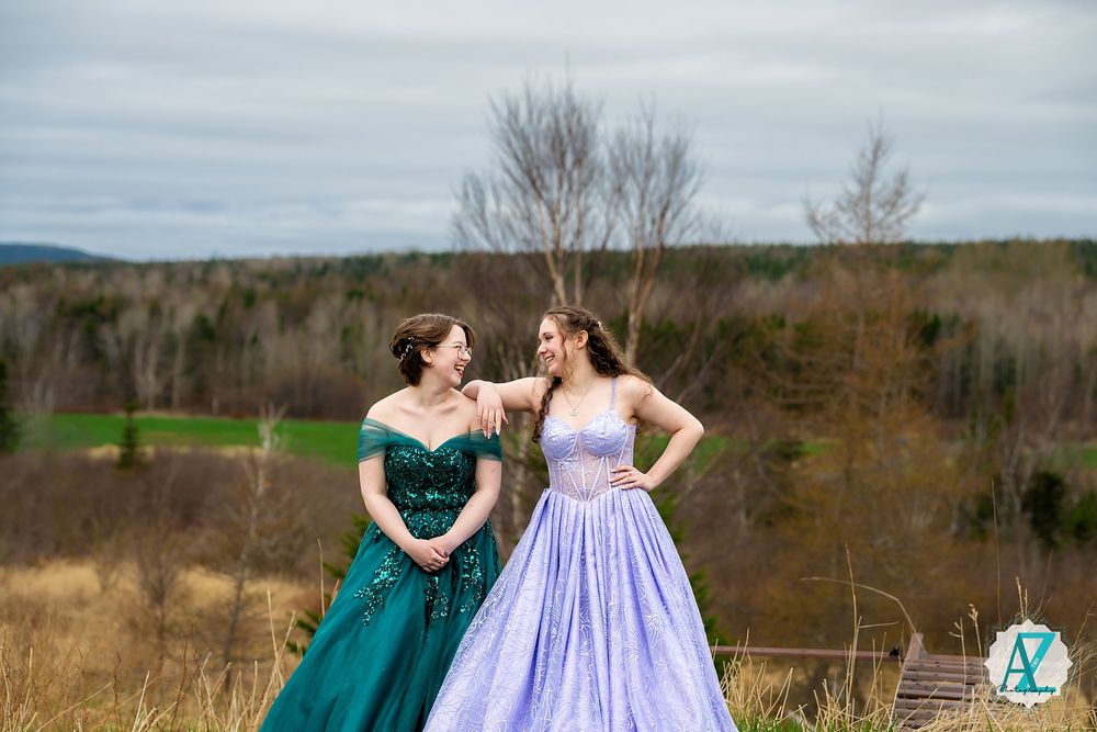 Two people in formal ballgowns, one emerald green and one lavender, stand together on a hilltop overlooking autumn landscape.