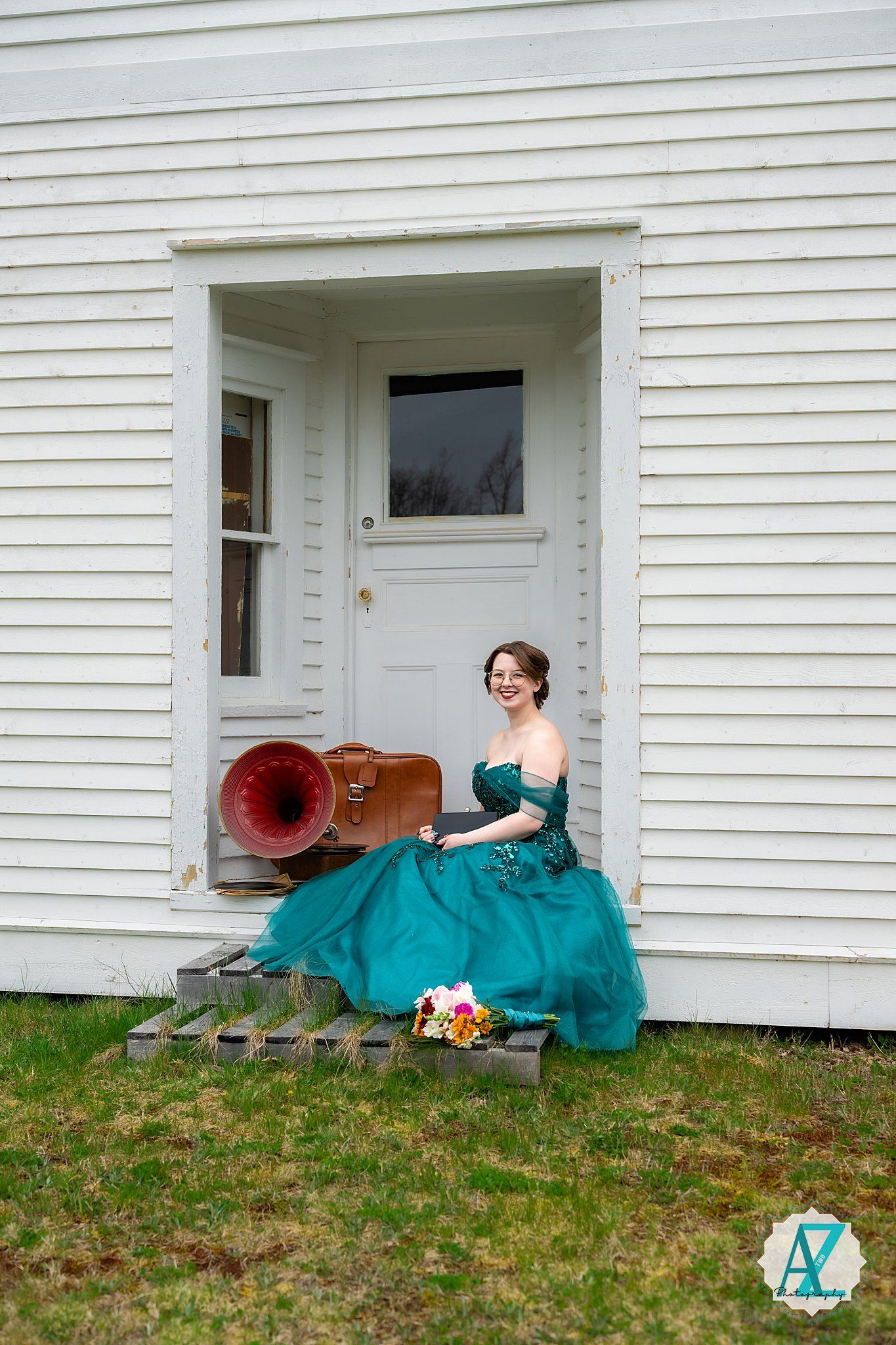 A person in a teal ball gown sits on wooden steps by a white cottage door with a vintage red phonograph nearby.