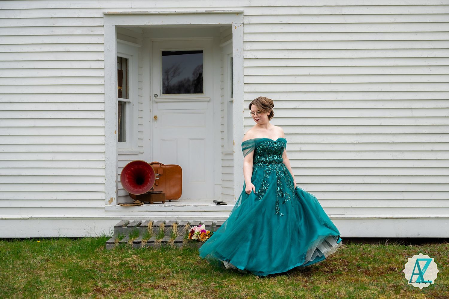 A person in a flowing teal formal gown twirls in front of a white clapboard house with a red speaker on the porch.