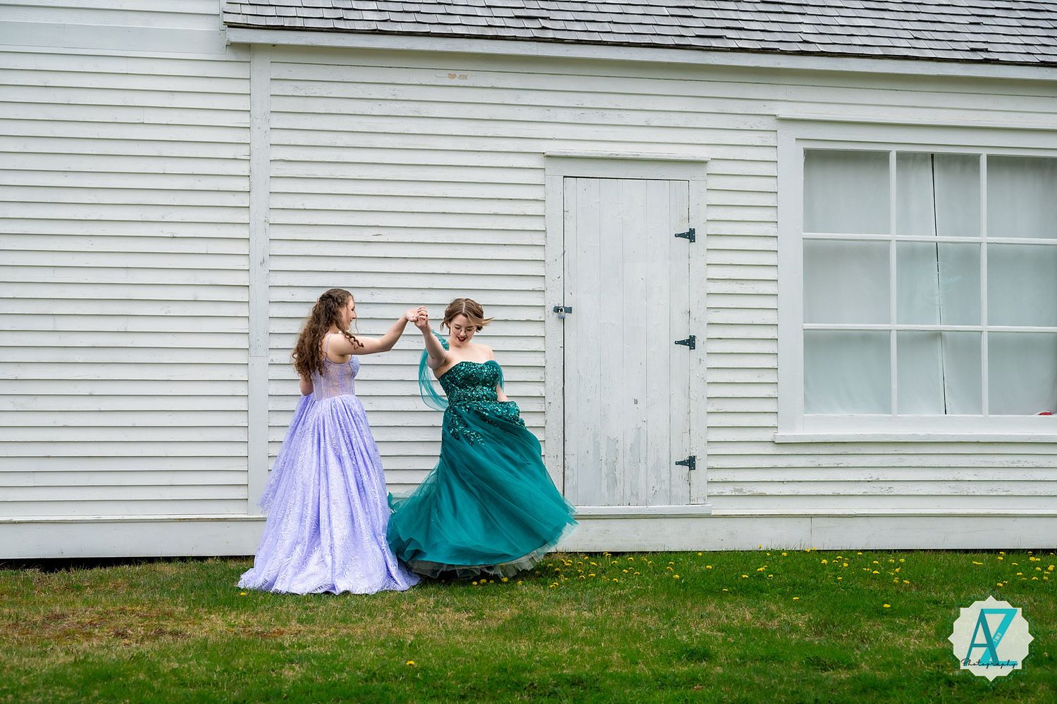 Two figures in flowing formal dresses, one purple and one teal, dancing together joyfully on a lawn in front of a white building.