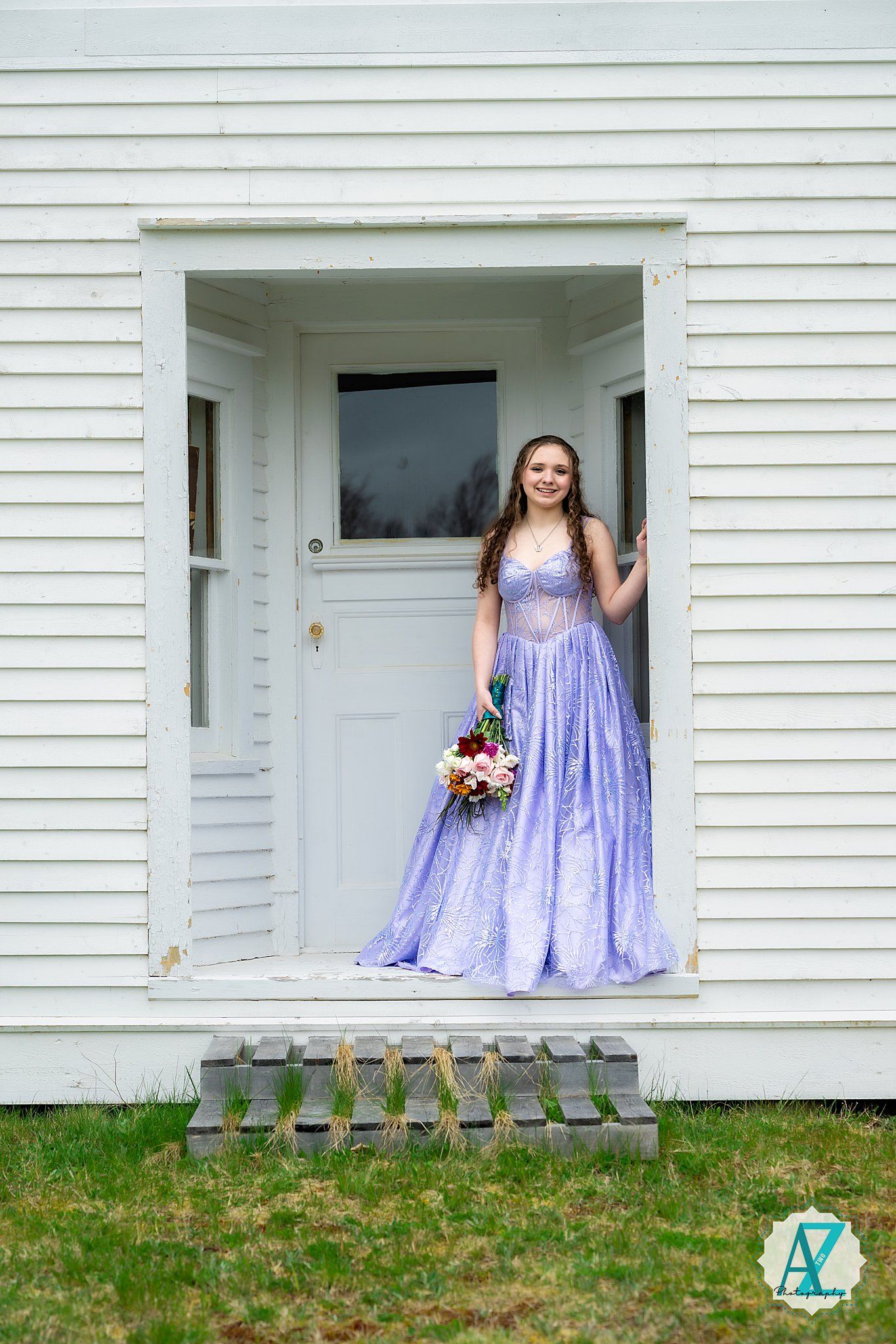 A figure in a flowing lavender prom dress poses in a white doorway of a classic clapboard house.