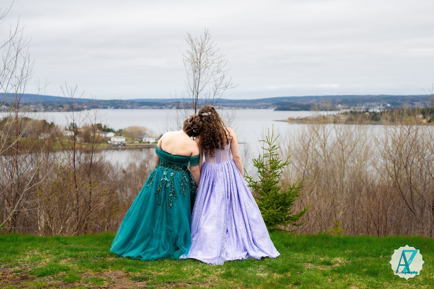 Two people in elegant ball gowns stand together admiring the scenic lake view from a hilltop.