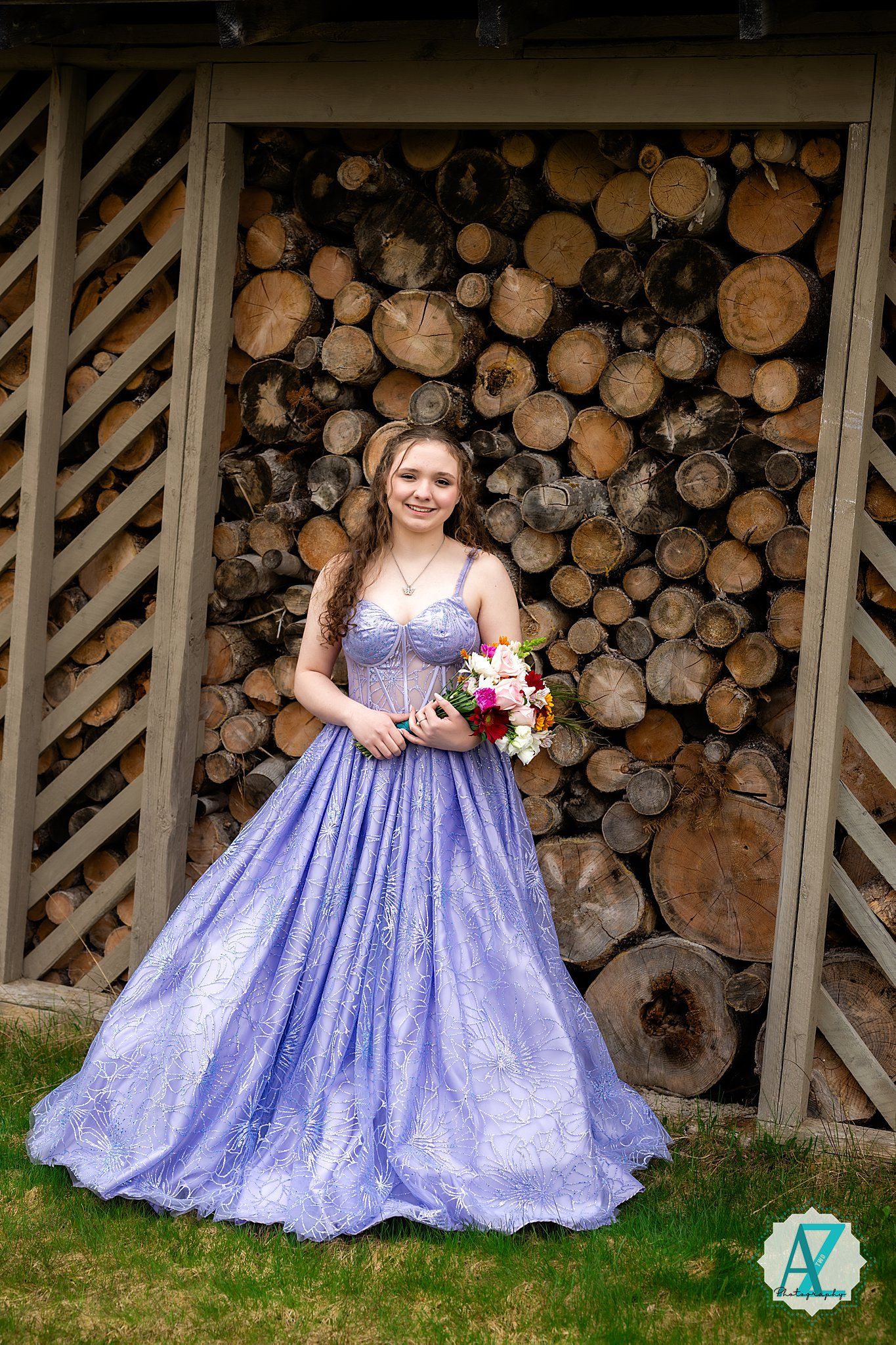A person in a lavender ballgown poses with flowers in front of stacked firewood at a rustic venue.