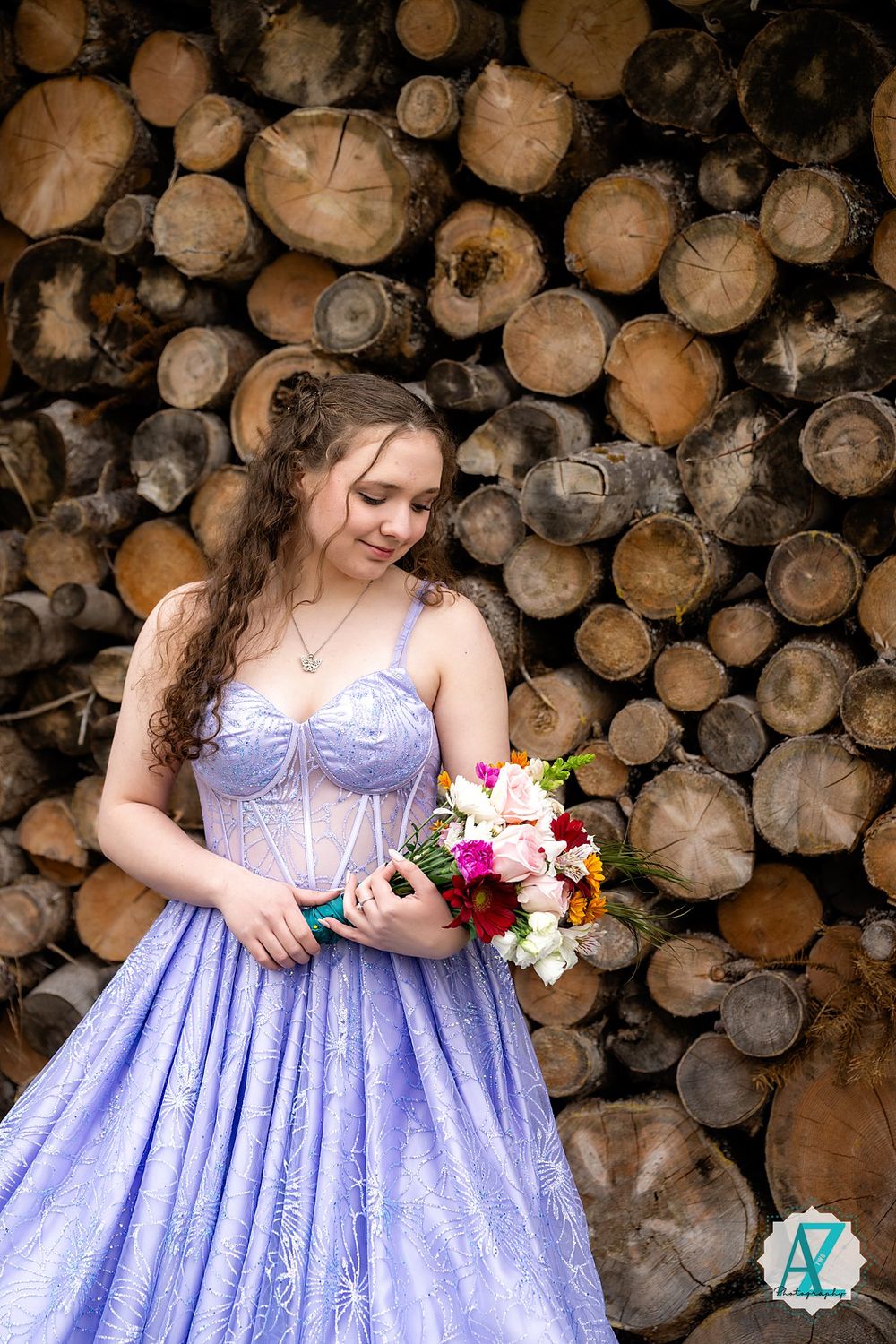 A person in a purple gown holds a bouquet while posing against a backdrop of stacked wooden logs.