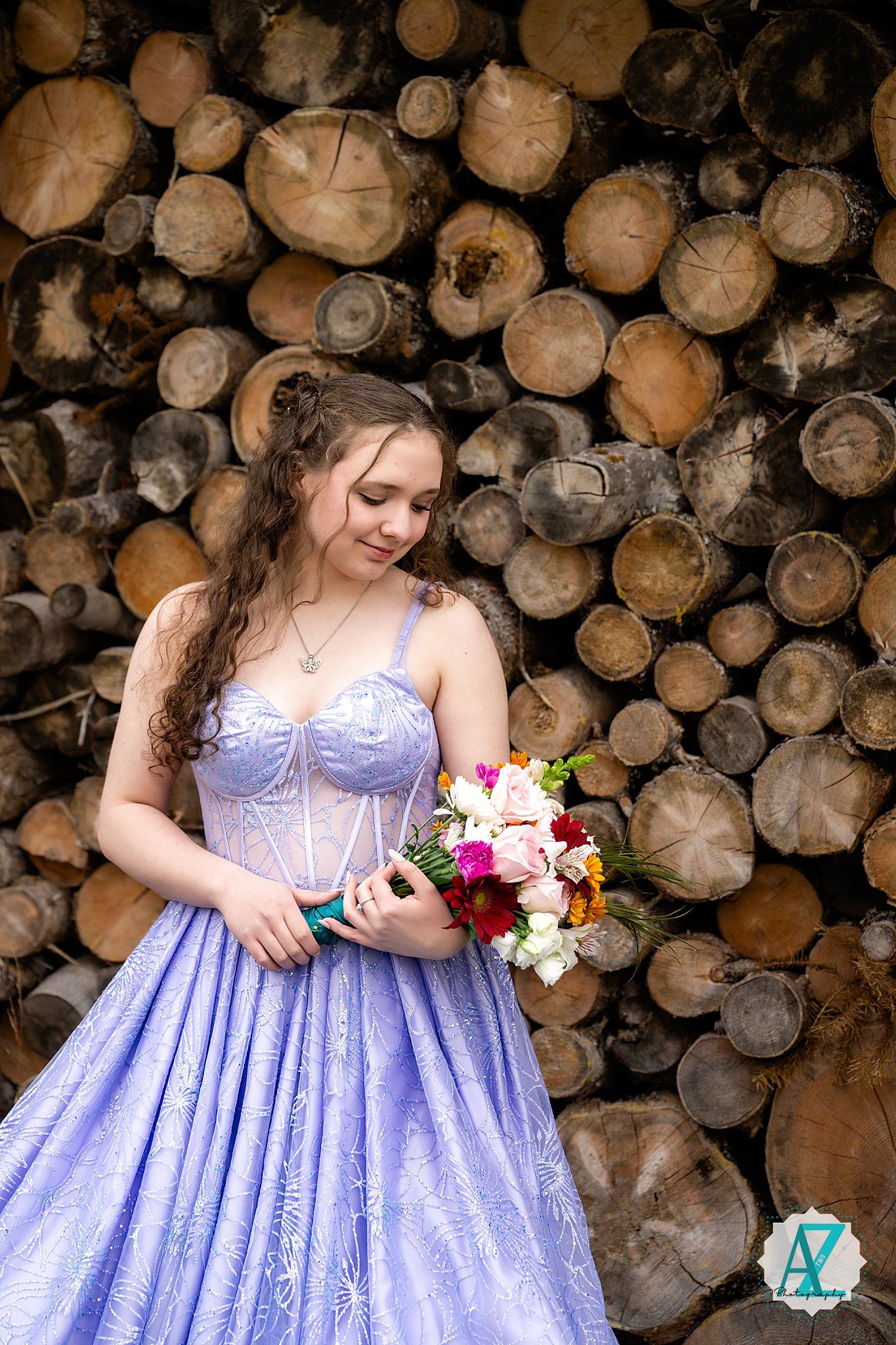 A person in a purple gown holds a bouquet while posing against a backdrop of stacked wooden logs.