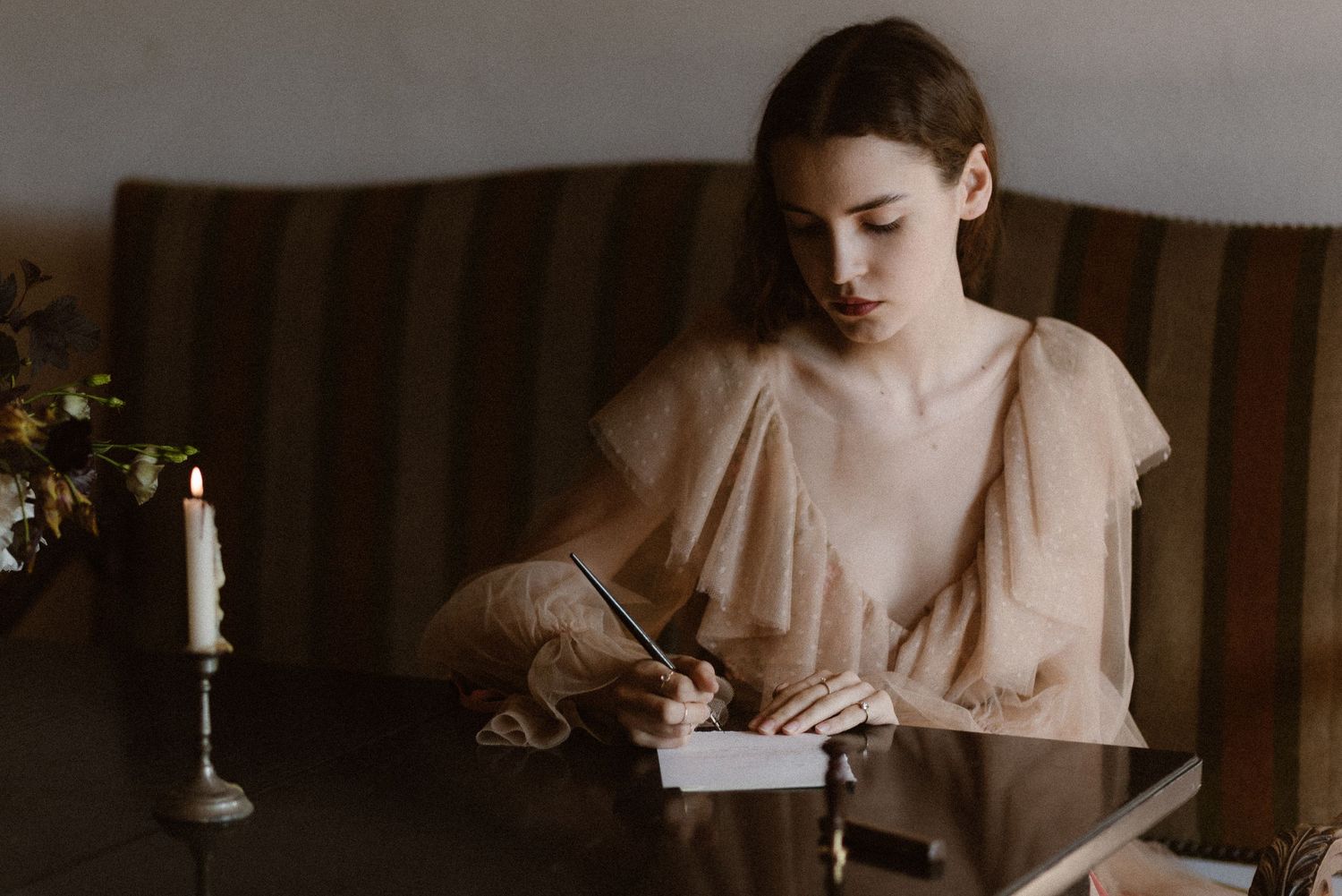 A person in a vintage-style romantic beige dress writes at a wooden table with a candlestick nearby in moody lighting.