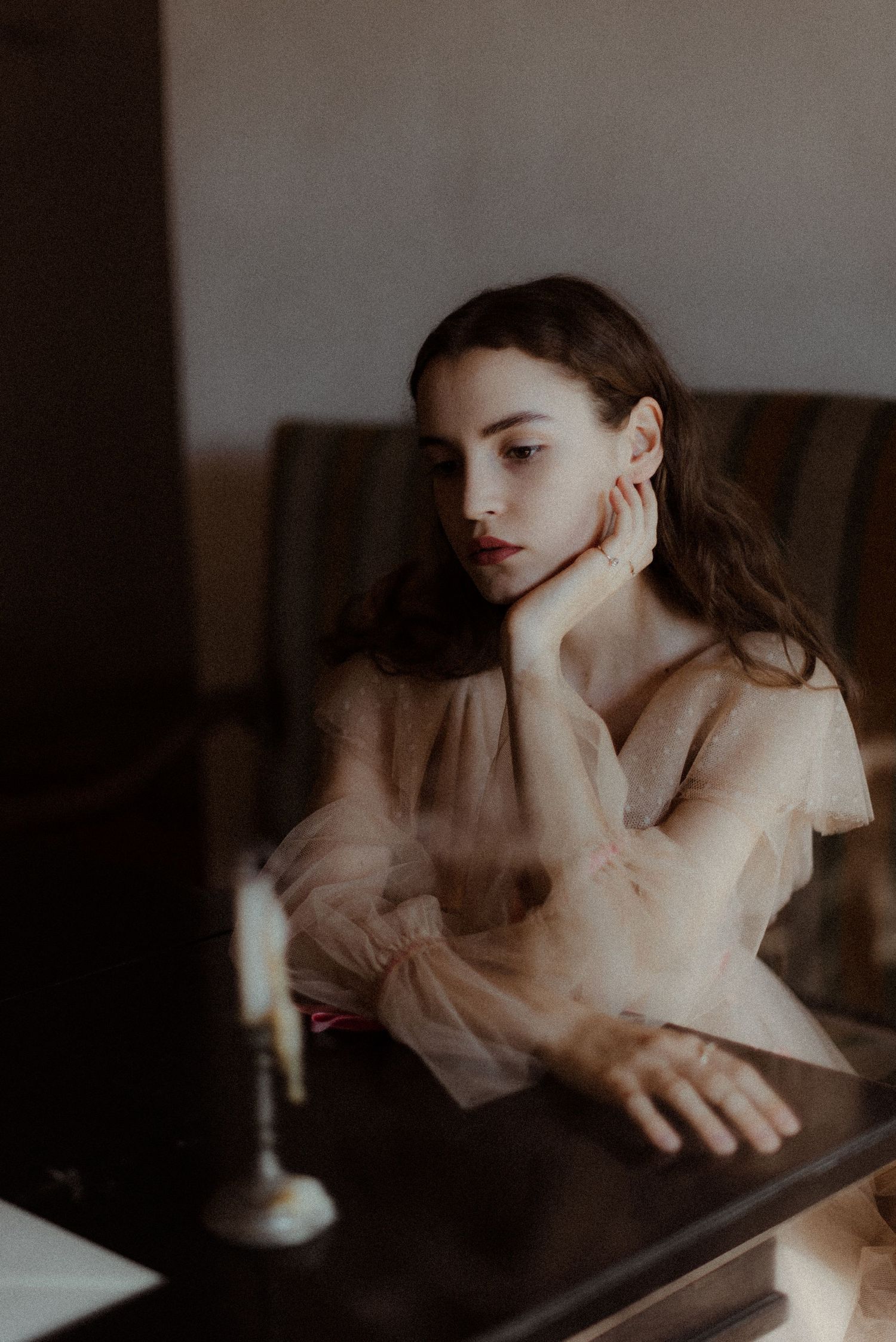 Artistic portrait in moody lighting shows figure in sheer blouse looking contemplative at table with single lit candle.