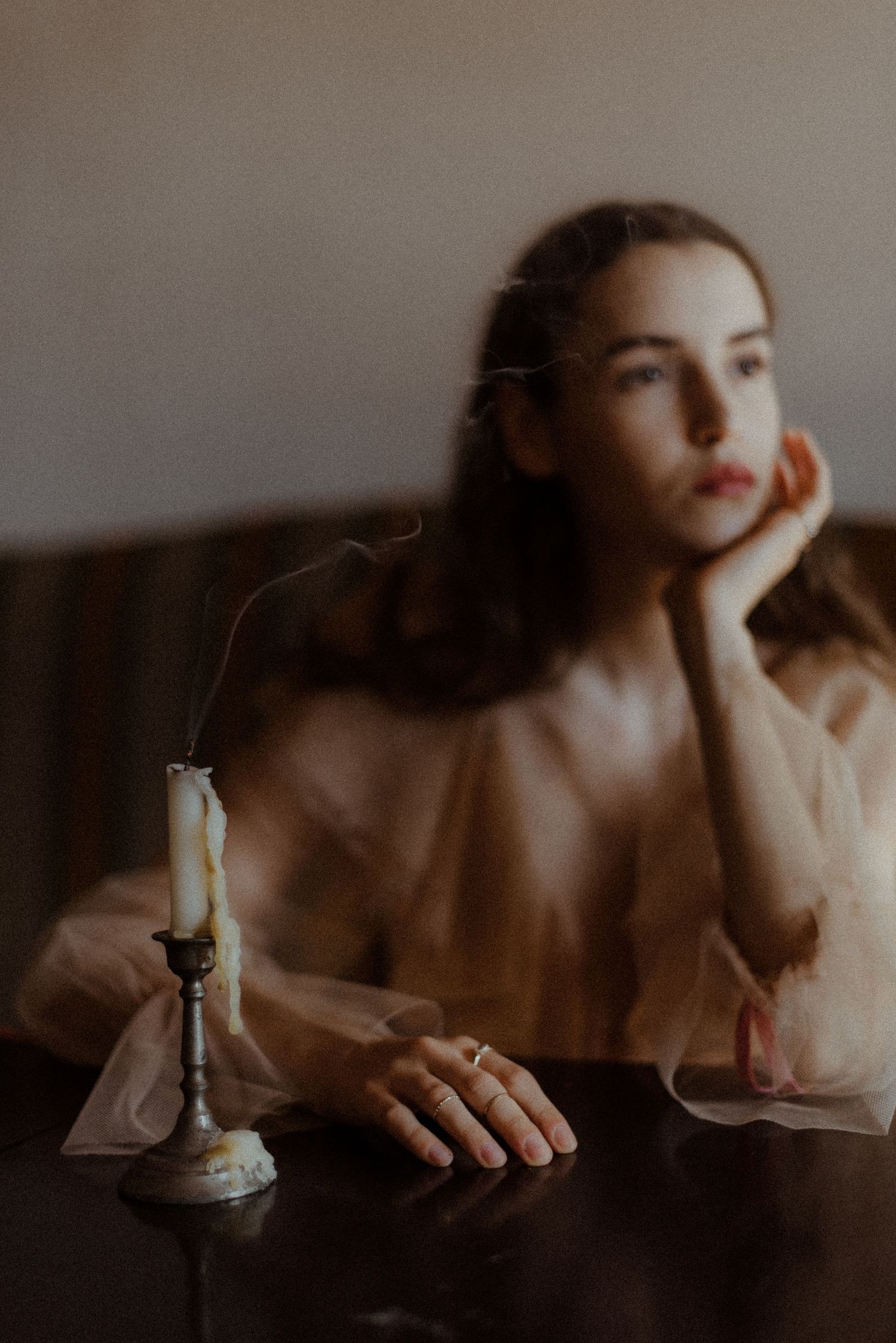 Moody vintage-style portrait of a person in sheer fabric sitting at table with lit candle in dark atmospheric setting.