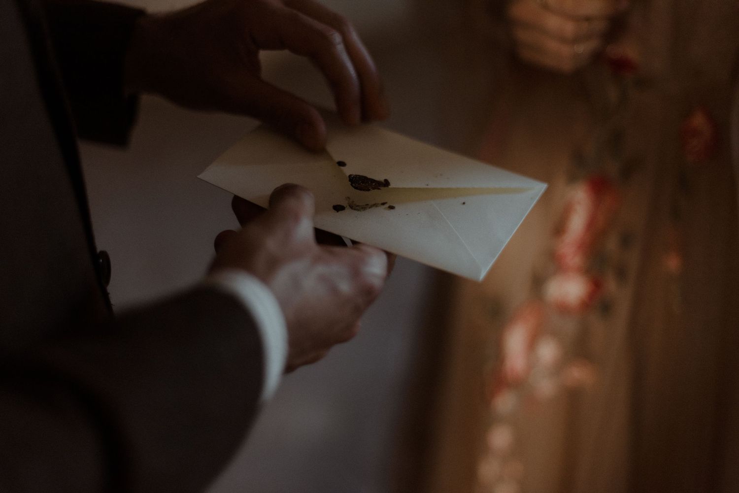 Hands in formal attire exchanging an envelope in a dimly lit setting with warm glowing lights in background.