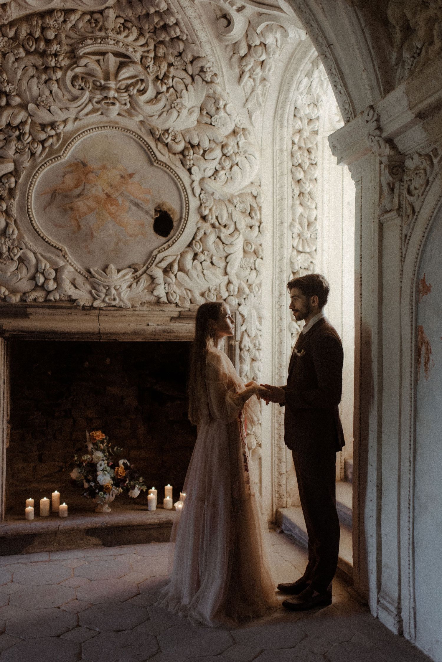 Couple standing in vintage room with ornate baroque wall carvings, fireplace with candles and elegant flower arrangement.