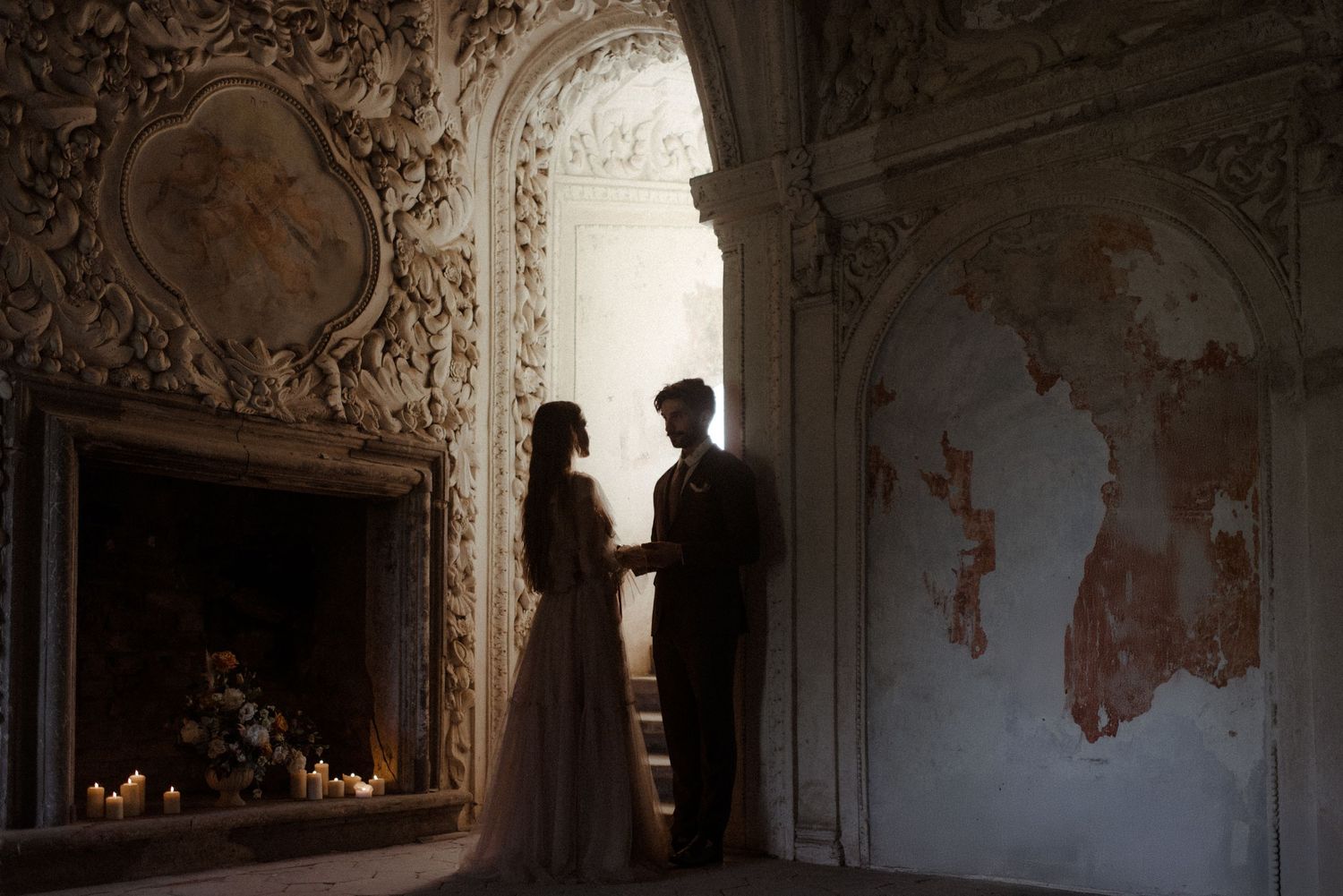 Two silhouettes stand intimately in a beautiful ornate room with a vintage fireplace and distressed wall details.