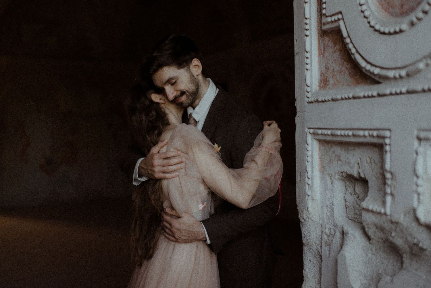 A romantic couple embraces in vintage formal attire as they dance in a dimly lit room with ornate white wall molding.