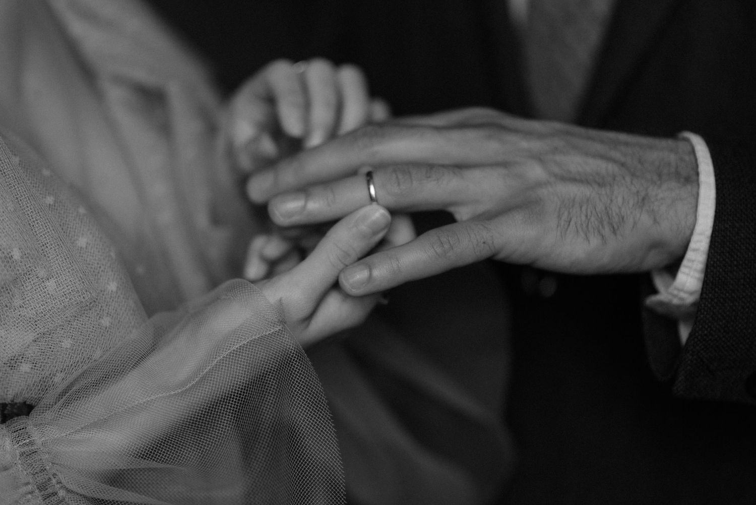 Black and white closeup of hands exchanging wedding rings during a marriage ceremony.