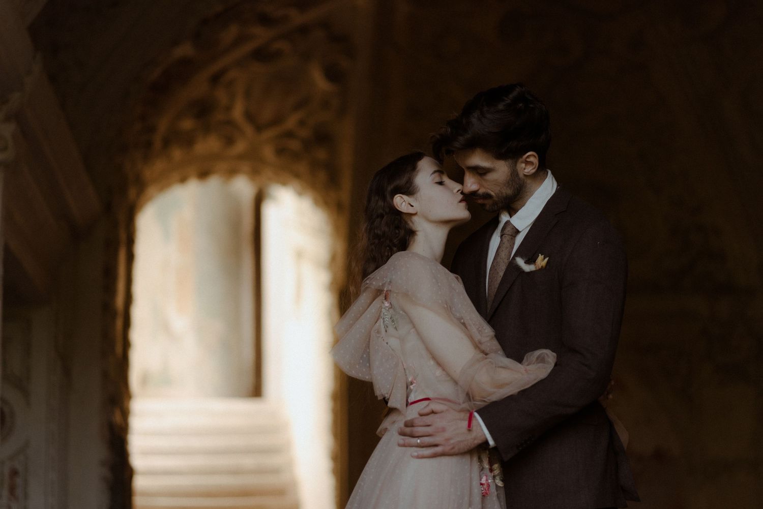 A romantic couple embraces in an elegant stone archway with soft lighting creating a dreamy atmosphere.