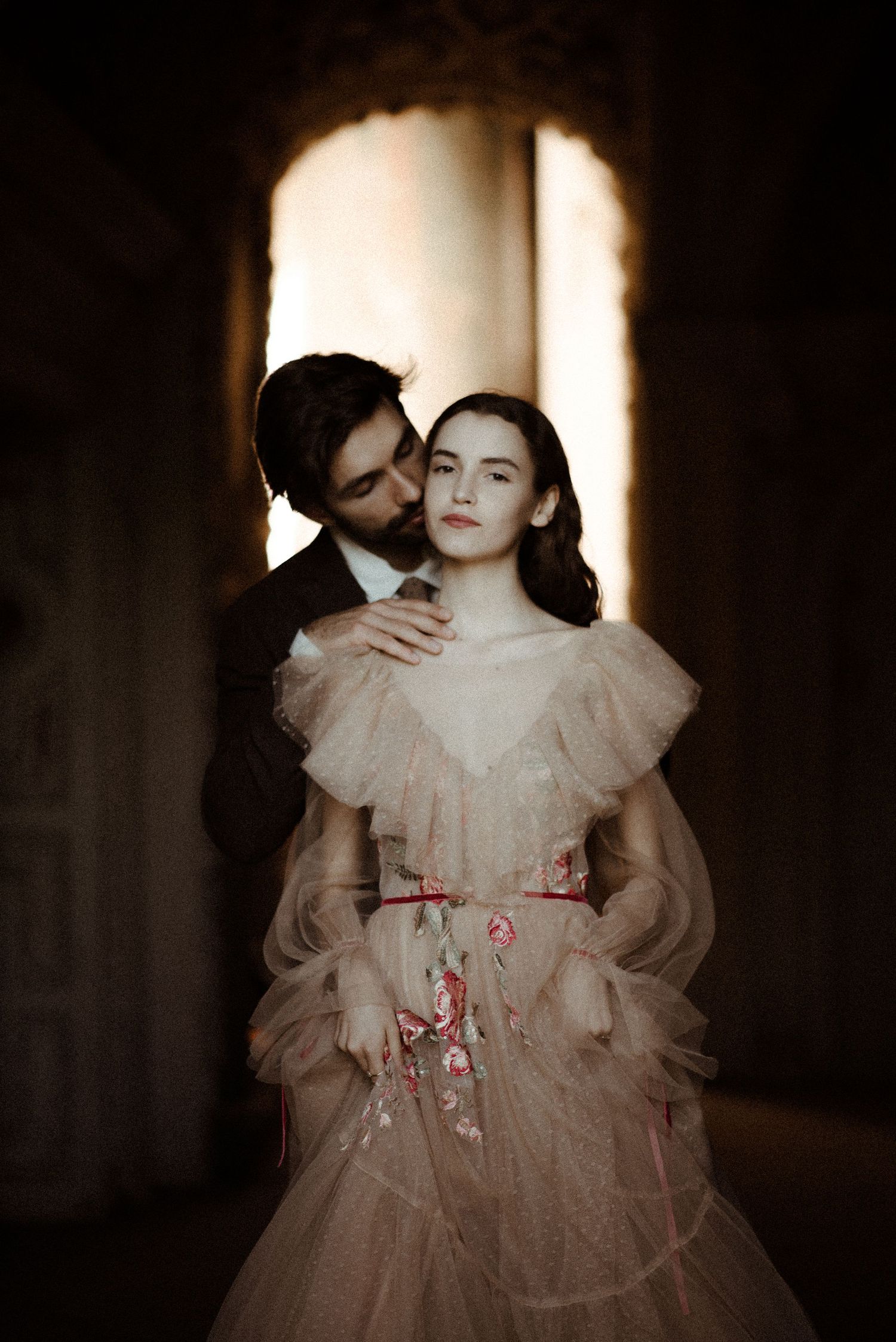 A romantic vintage-style portrait of a couple in formal wear with tulle and embroidered details in moody lighting.