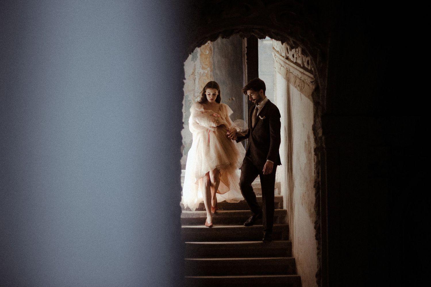 A couple in vintage attire descend stone stairs in a dimly lit historic building during a romantic photoshoot.