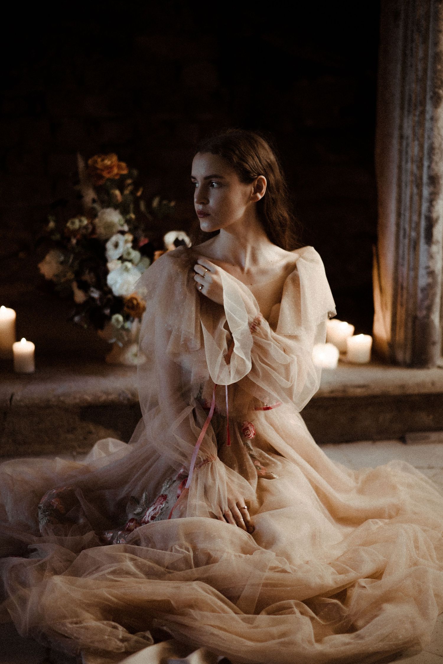 A romantic portrait in candlelight features a flowing champagne tulle gown and floral arrangements in a moody setting.