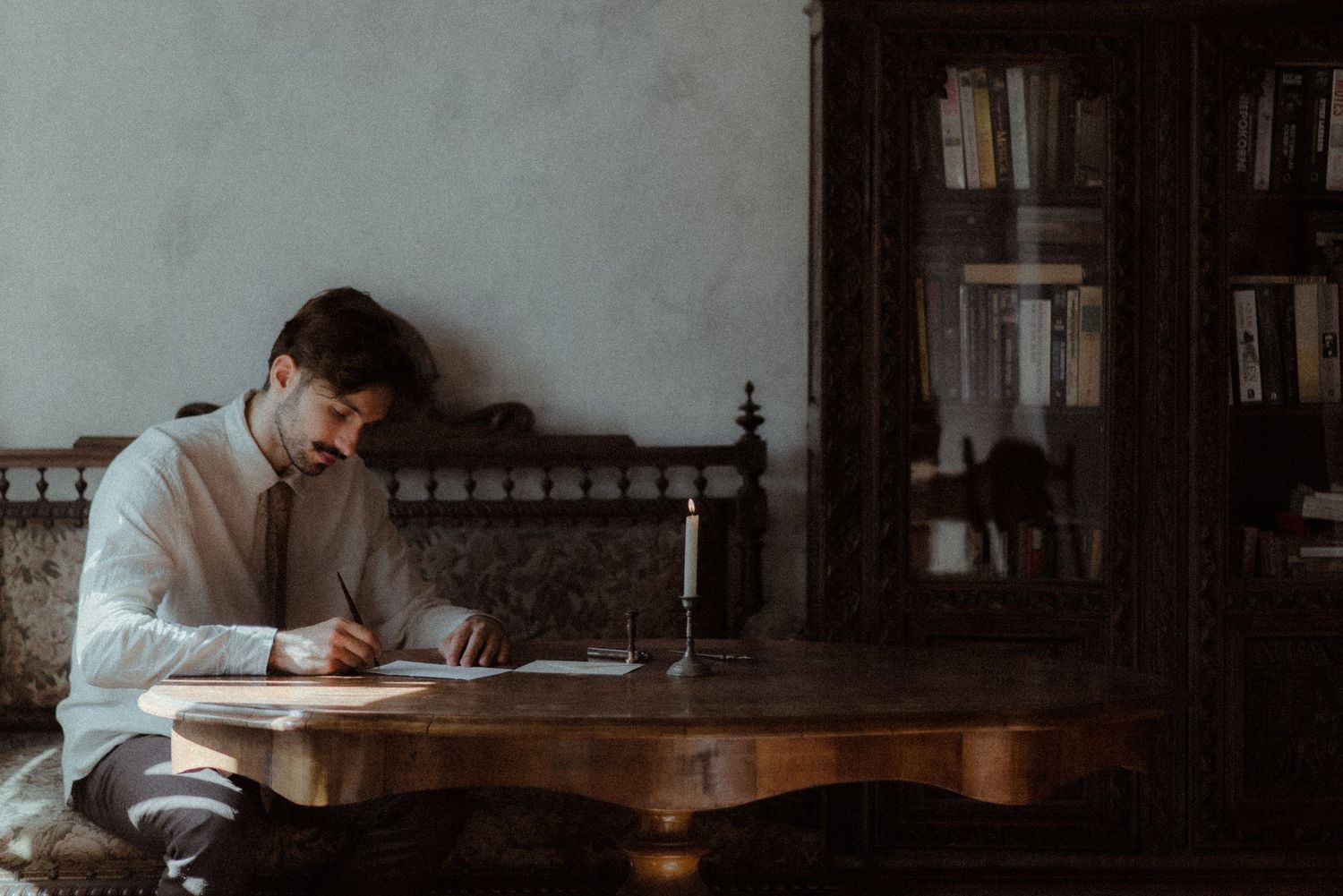 A person writes at an antique wooden desk near a bookshelf in a moody, atmospheric room.