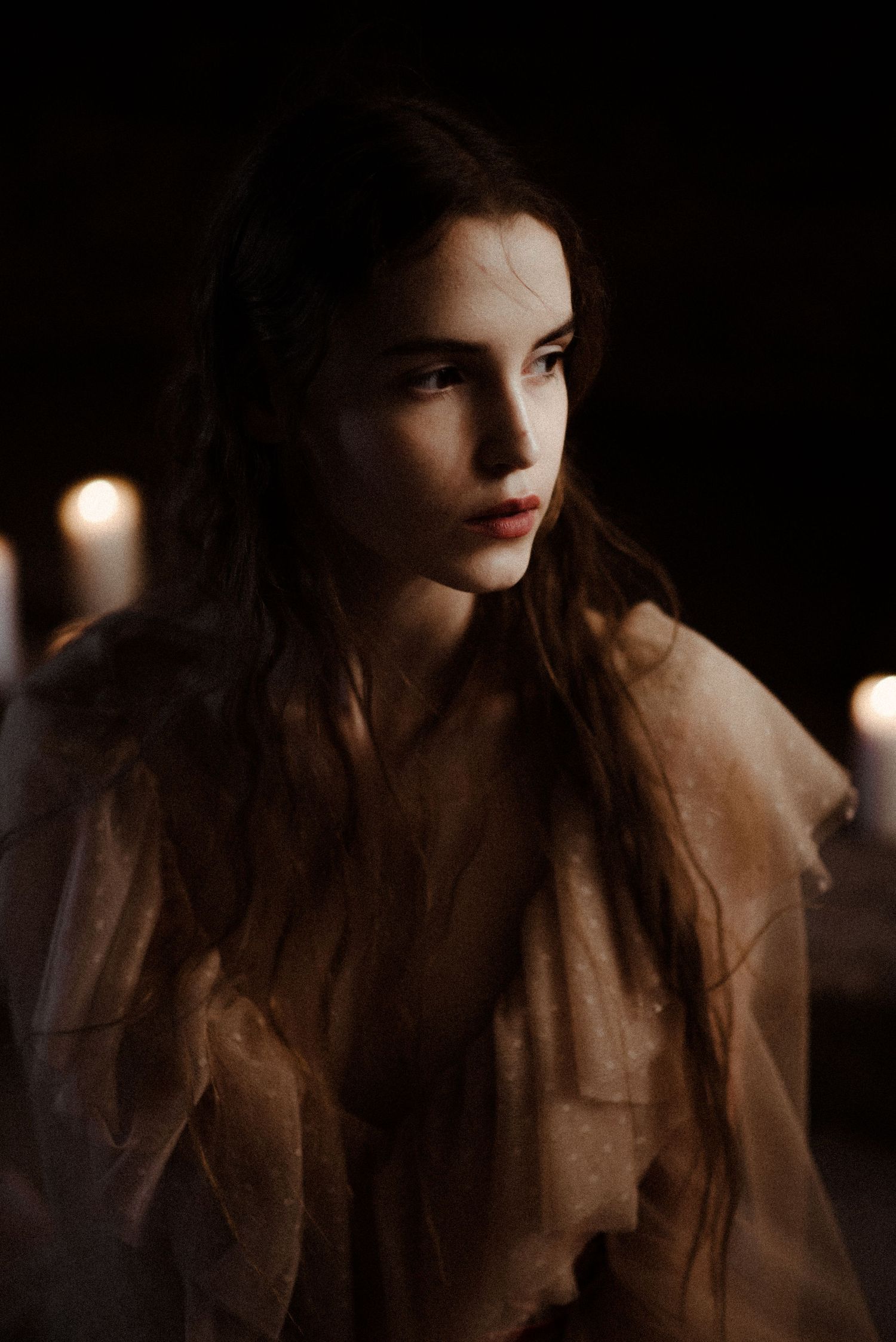 Moody atmospheric portrait with candlelight illuminating a soft brown dress against a dark background.