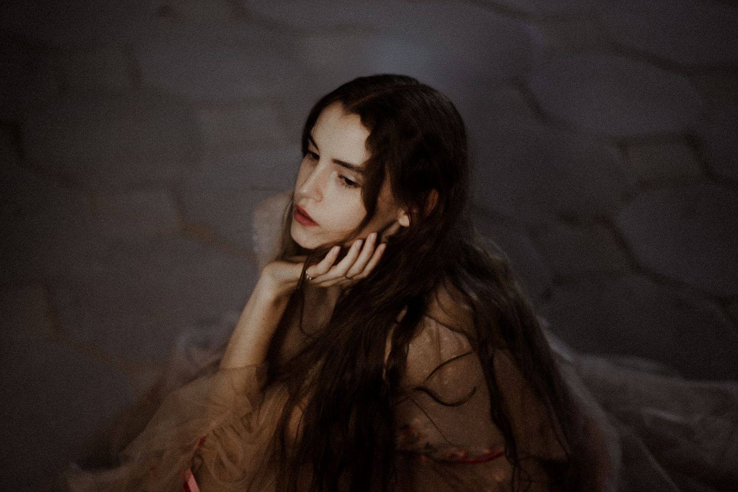 Moody portrait in dark lighting shows a dreamy figure with long dark hair resting their chin on hand.