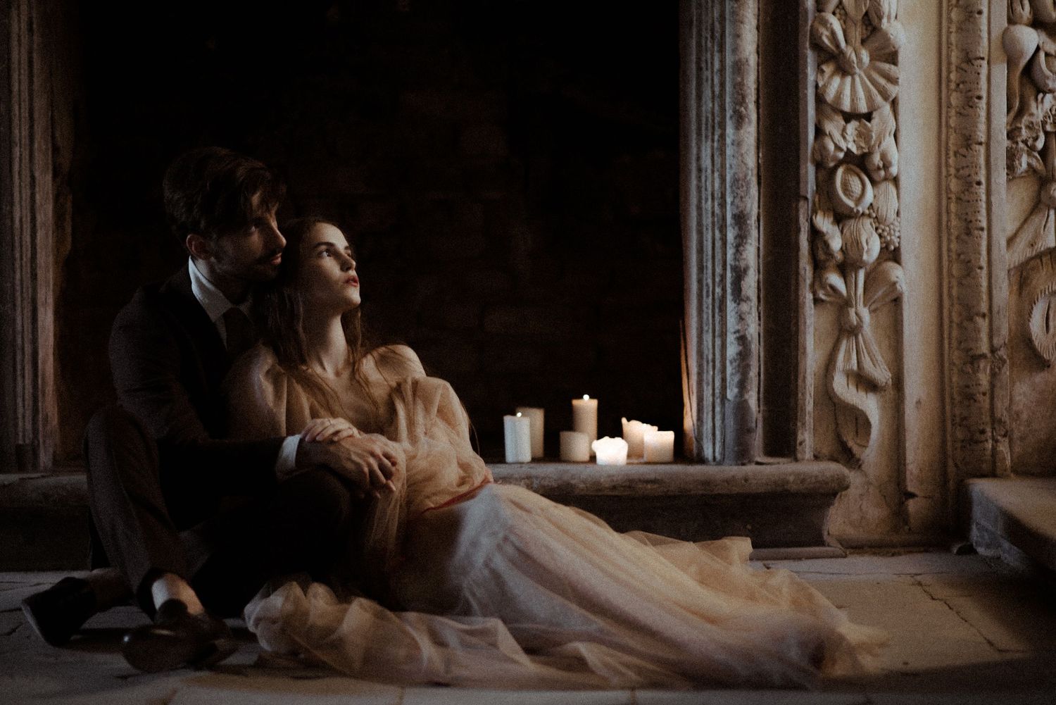 Romantic couple in formal attire sitting intimately by candlelight in an ornate vintage interior setting.