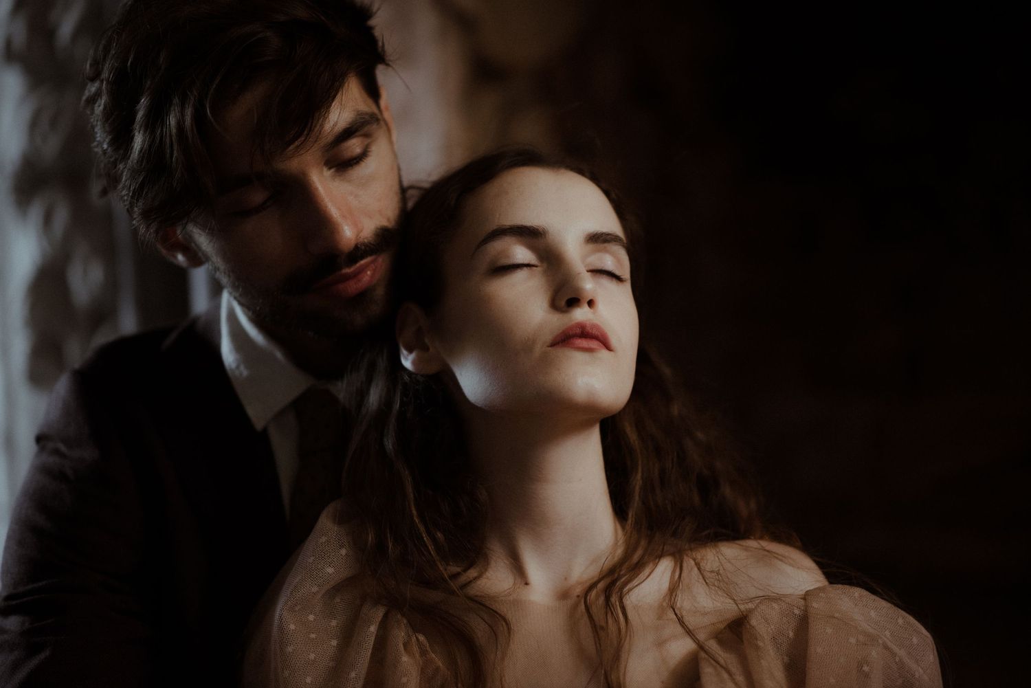 Dramatic moody couple portrait in dark tones with woman in brown dress tilting head back while partner leans close.