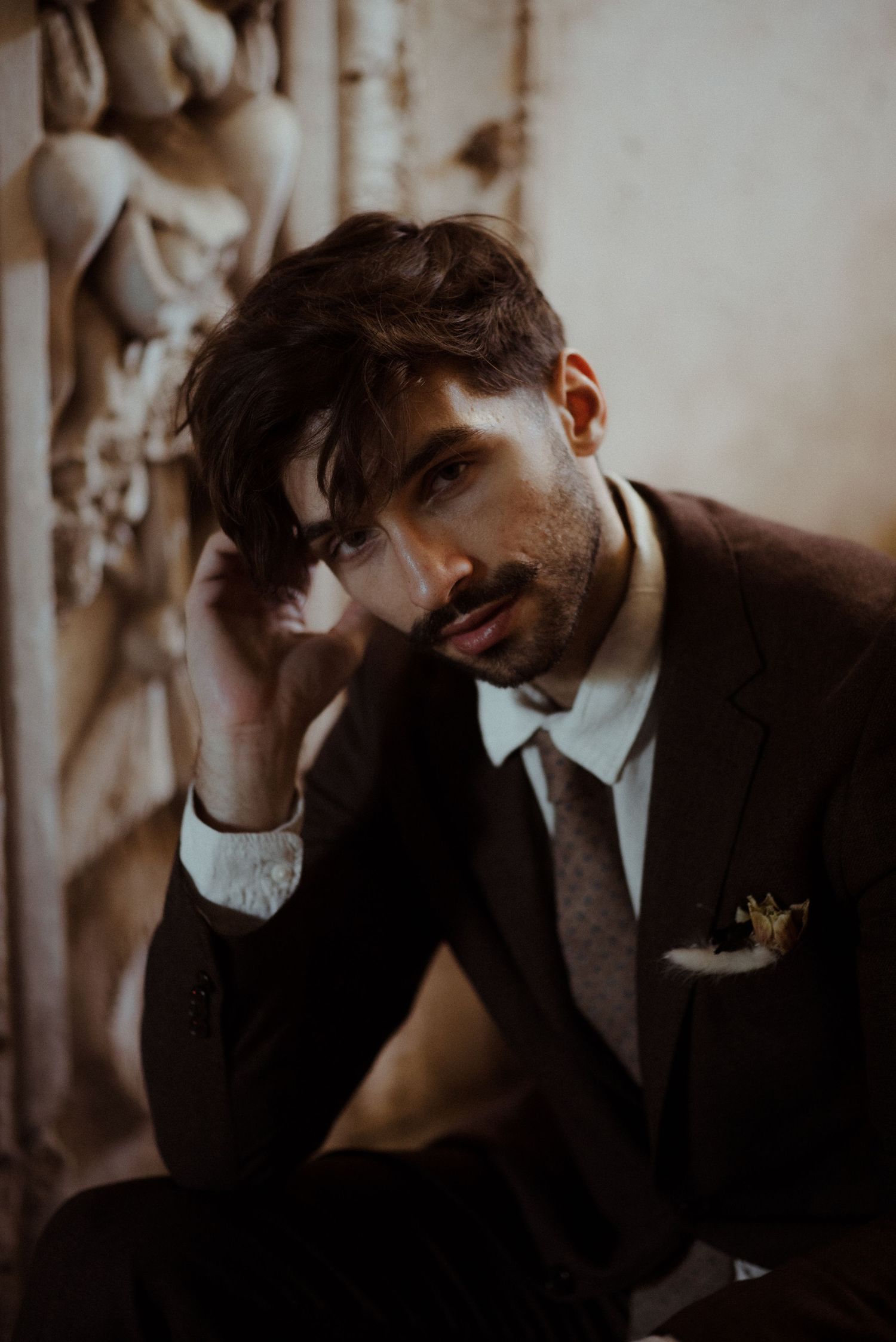 A moody portrait in brown tones shows someone in a suit and tie resting their hand against their head.