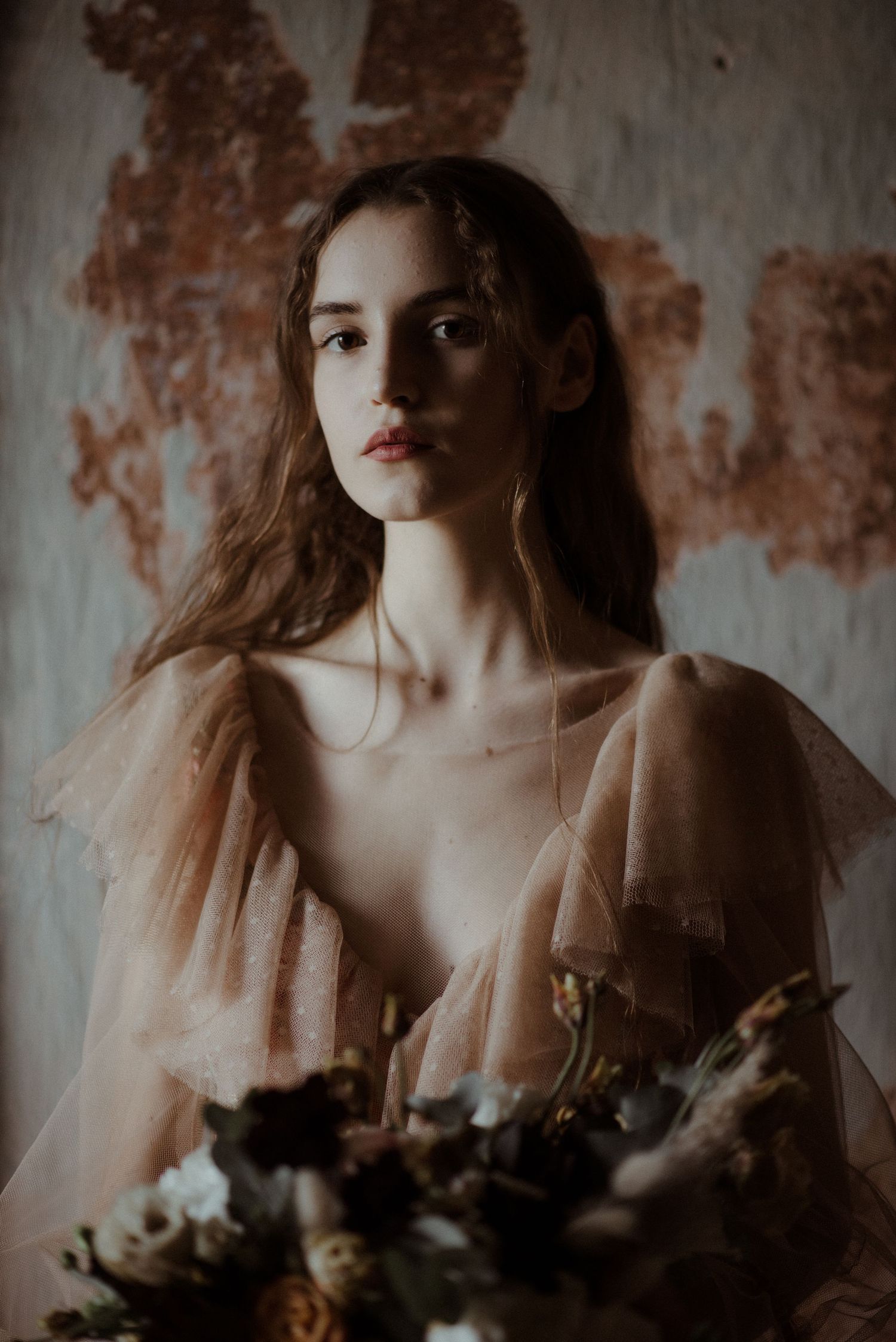 Moody fine art portrait with soft beige tones and floral arrangement against distressed textured wall background.