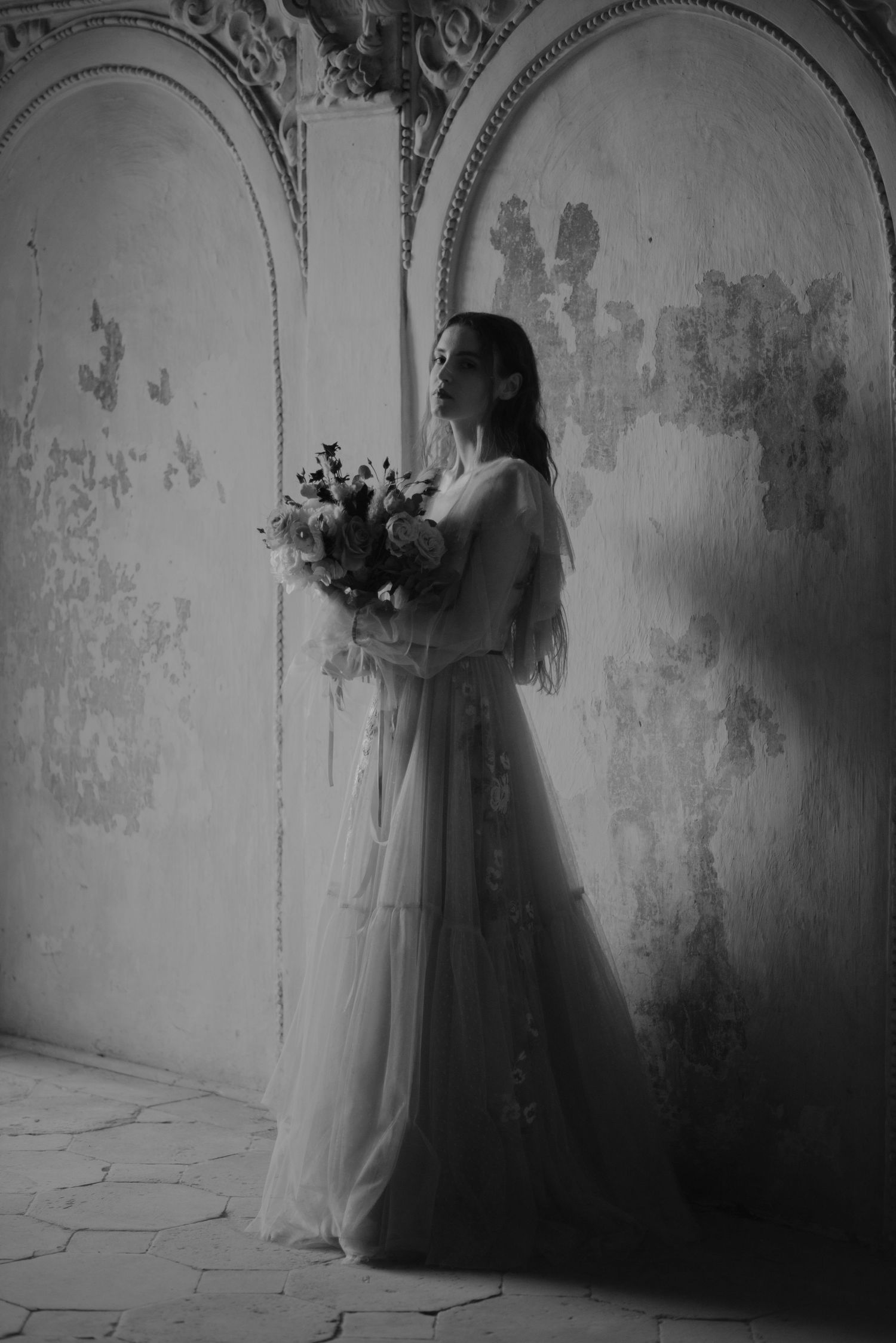 A romantic black and white photo of someone in a flowing wedding dress holding a bouquet against a vintage wall.