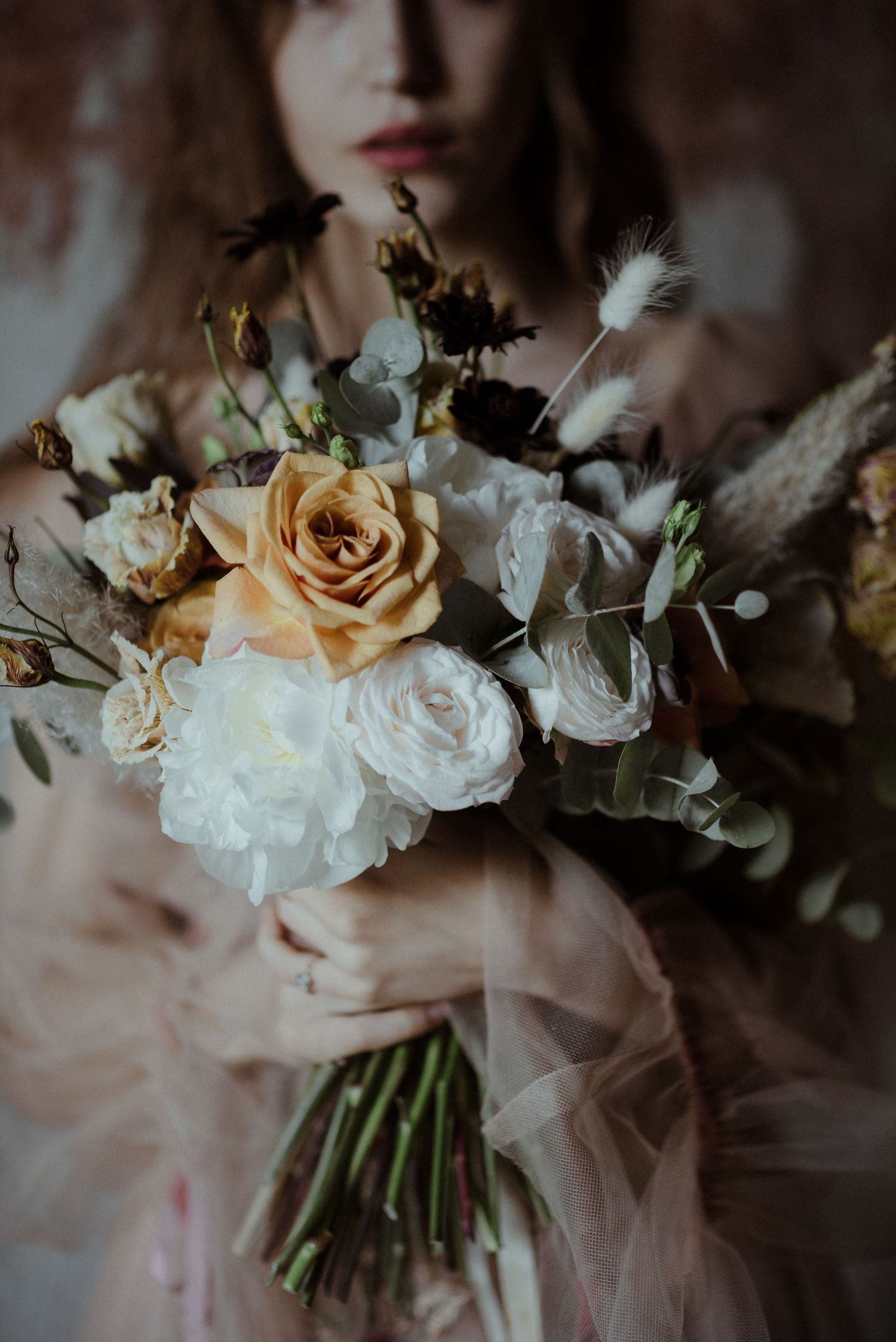 A romantic wedding bouquet with peach and white roses, wispy feathers and eucalyptus leaves against a moody dark background.
