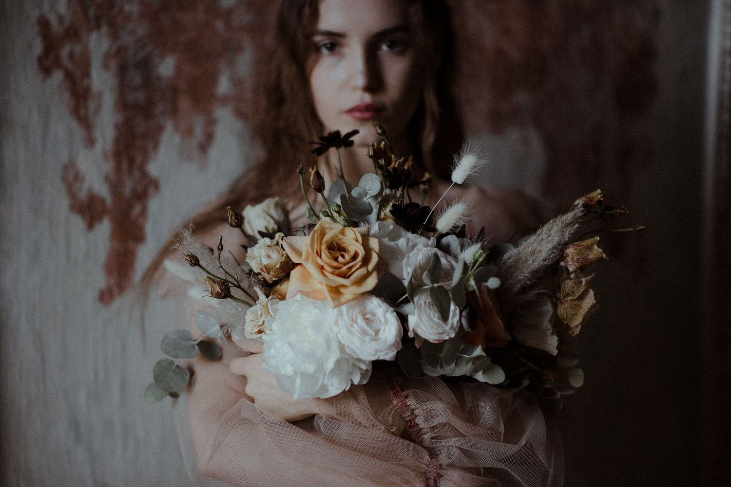 Person holding elegant autumn-toned floral bouquet with roses and dried elements against textured wall in moody lighting.